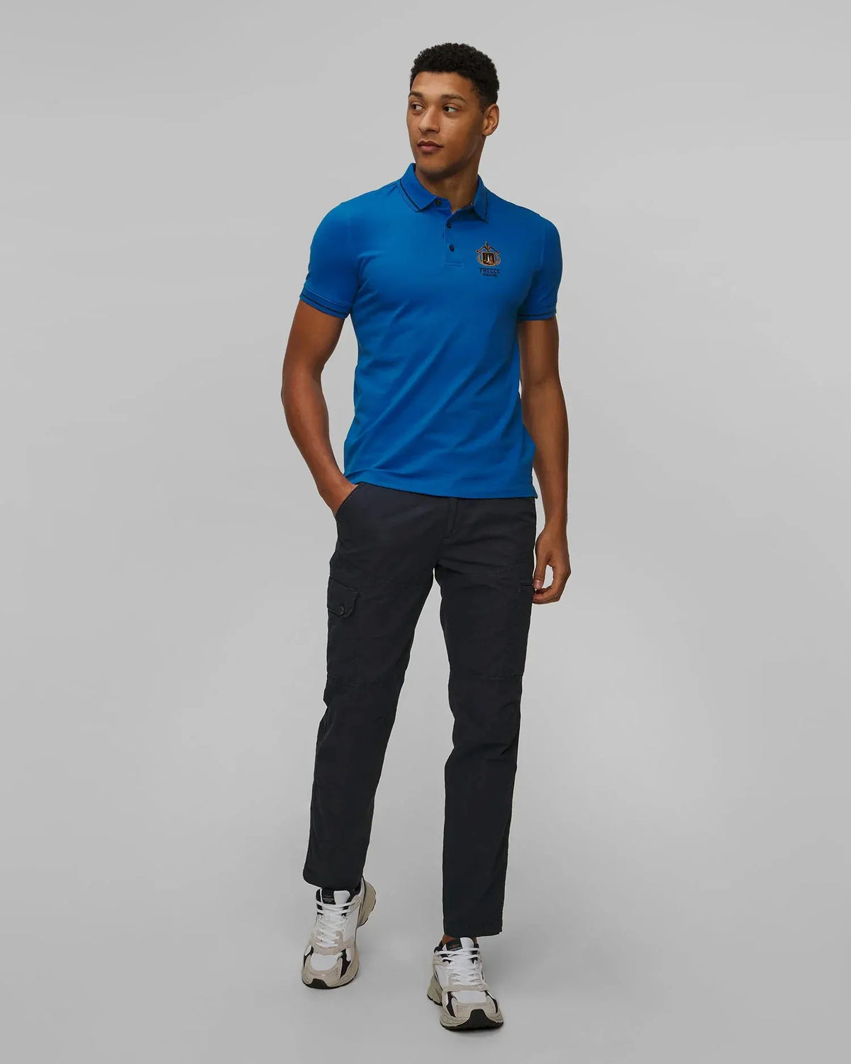 Modello uomo indossa polo blu con logo ricamato, pantaloni cargo neri e sneakers bianche.