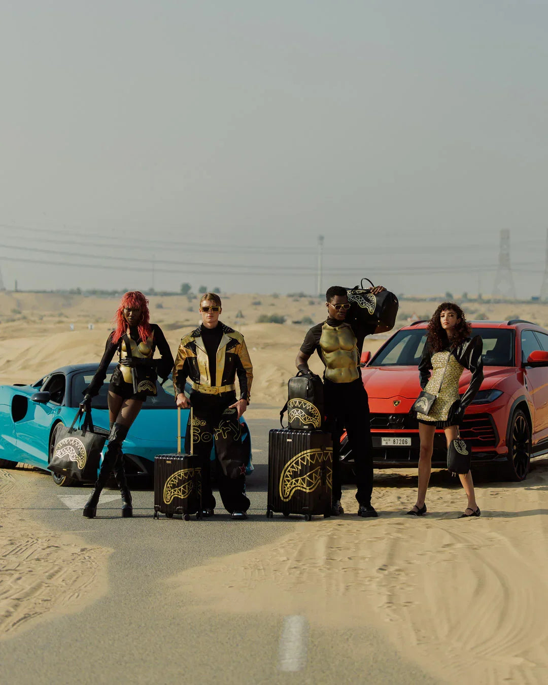 Quattro modelli posano in abbigliamento e accessori moda in pelle nera e oro, con valigie stampate, su una strada nel deserto davanti a supercar sportive.