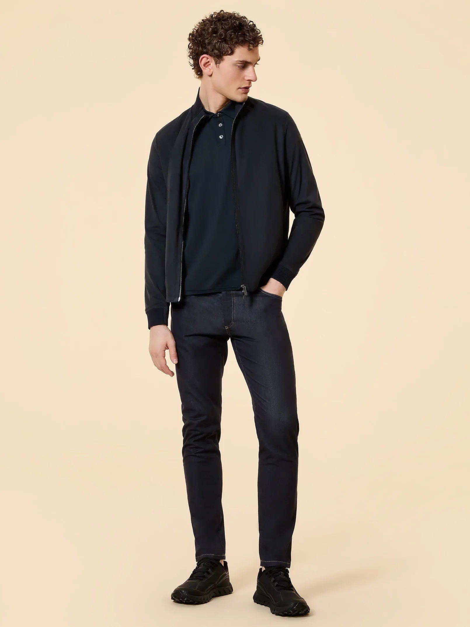 Uomo con giacca blu scuro, polo e jeans, look casual elegante su sfondo beige.