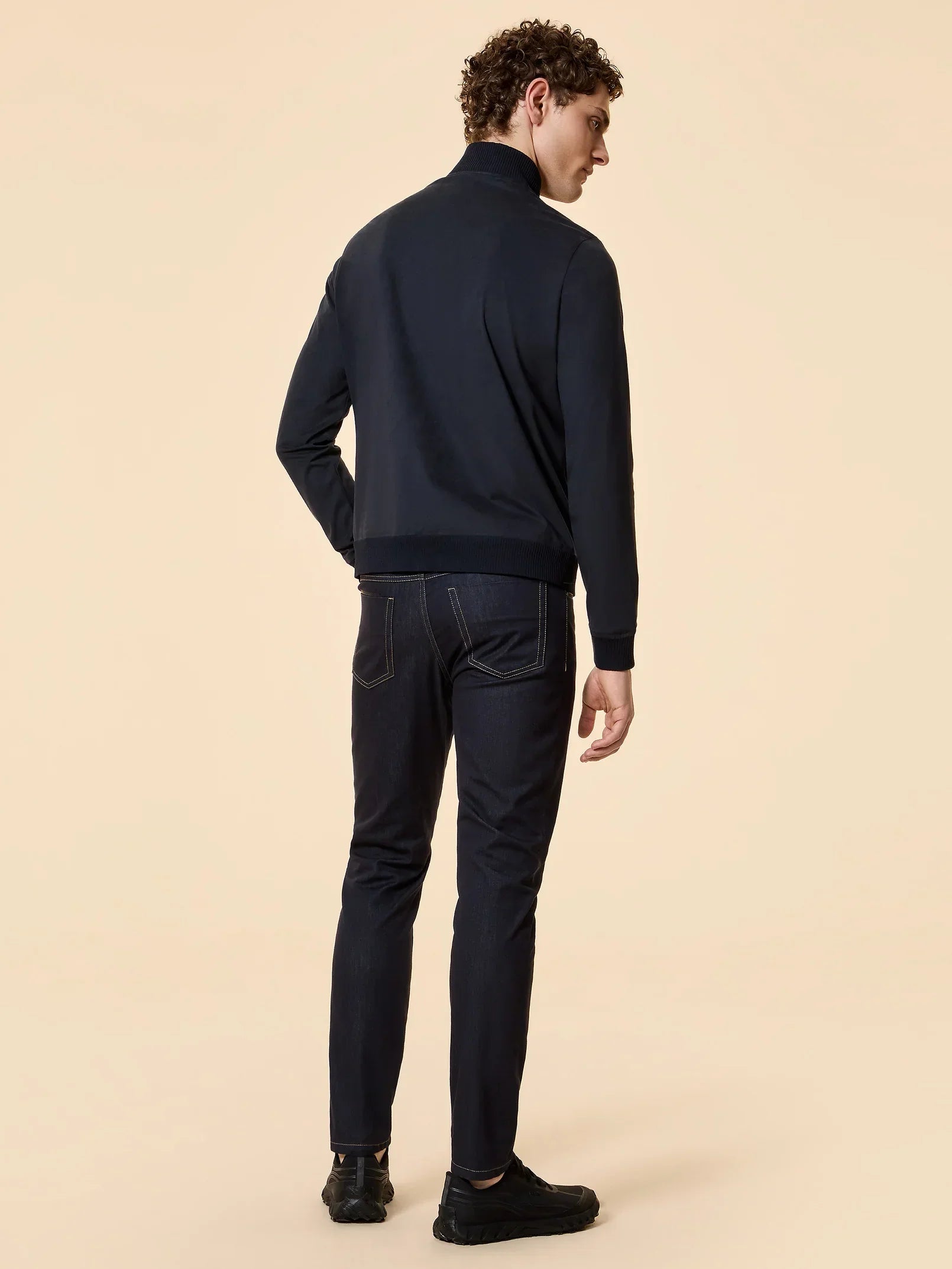 Modello uomo di spalle con giacca blu scuro, jeans slim e sneakers nere su sfondo beige.