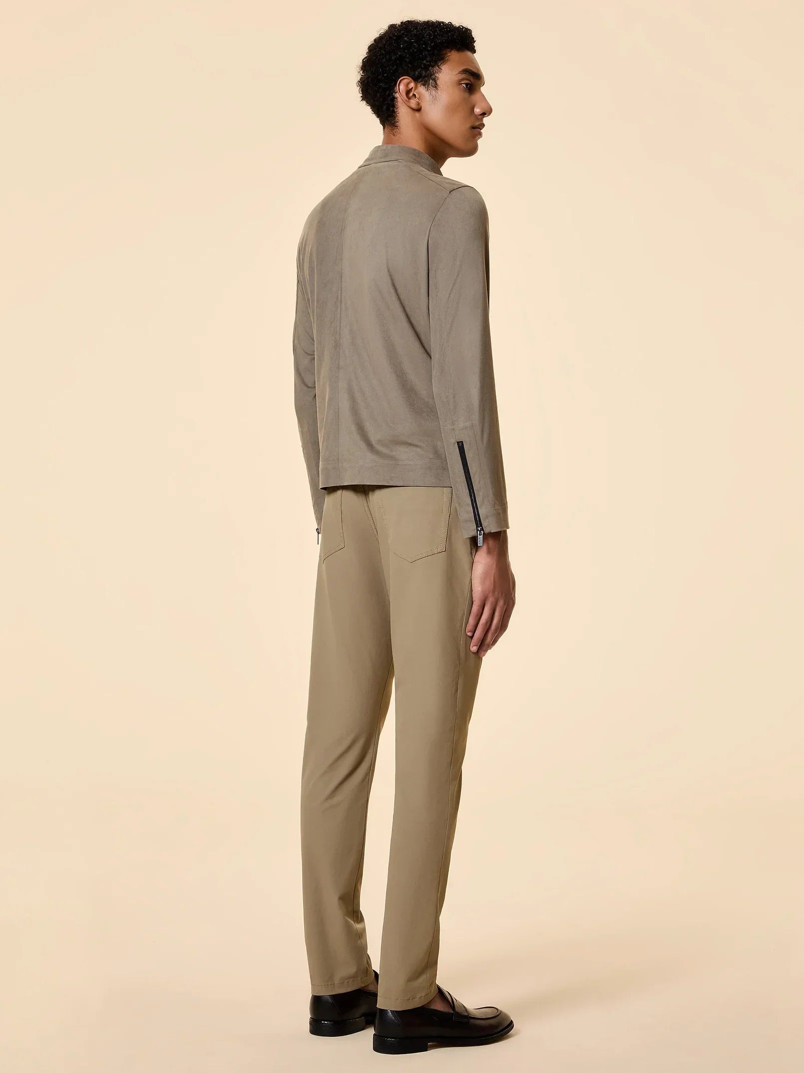 Modello uomo di spalle indossa giacca e pantaloni beige eleganti, scarpe nere, sfondo neutro.