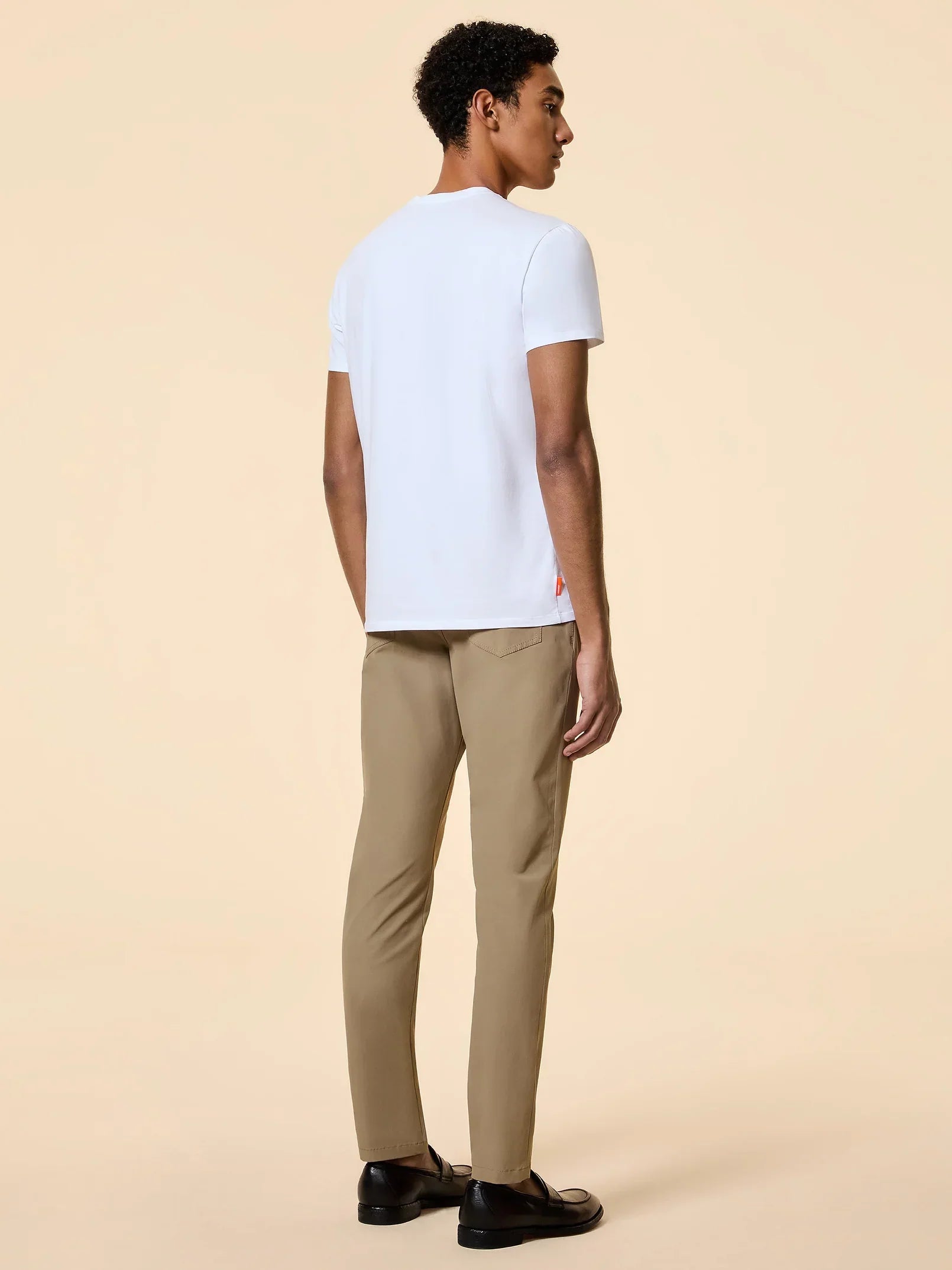 Uomo di spalle con t-shirt bianca, pantaloni beige e mocassini neri su sfondo neutro.
