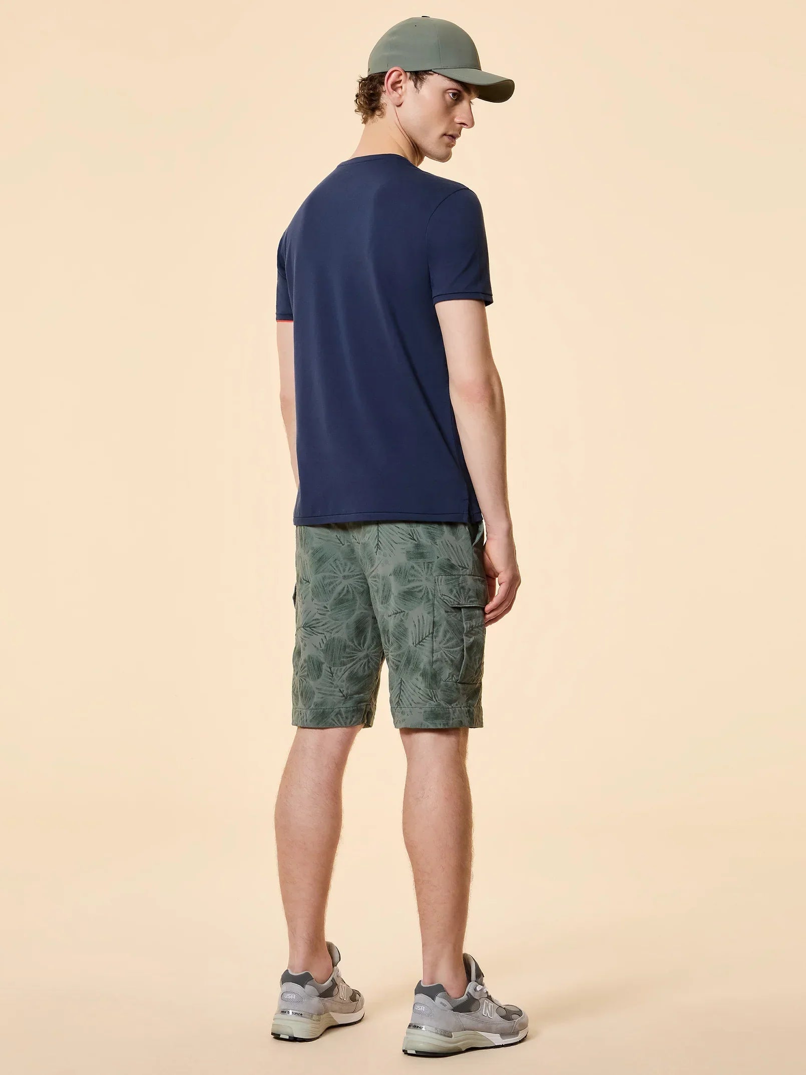 uomo di spalle con t-shirt blu, bermuda verdi a fantasia foglie, cappellino e sneakers