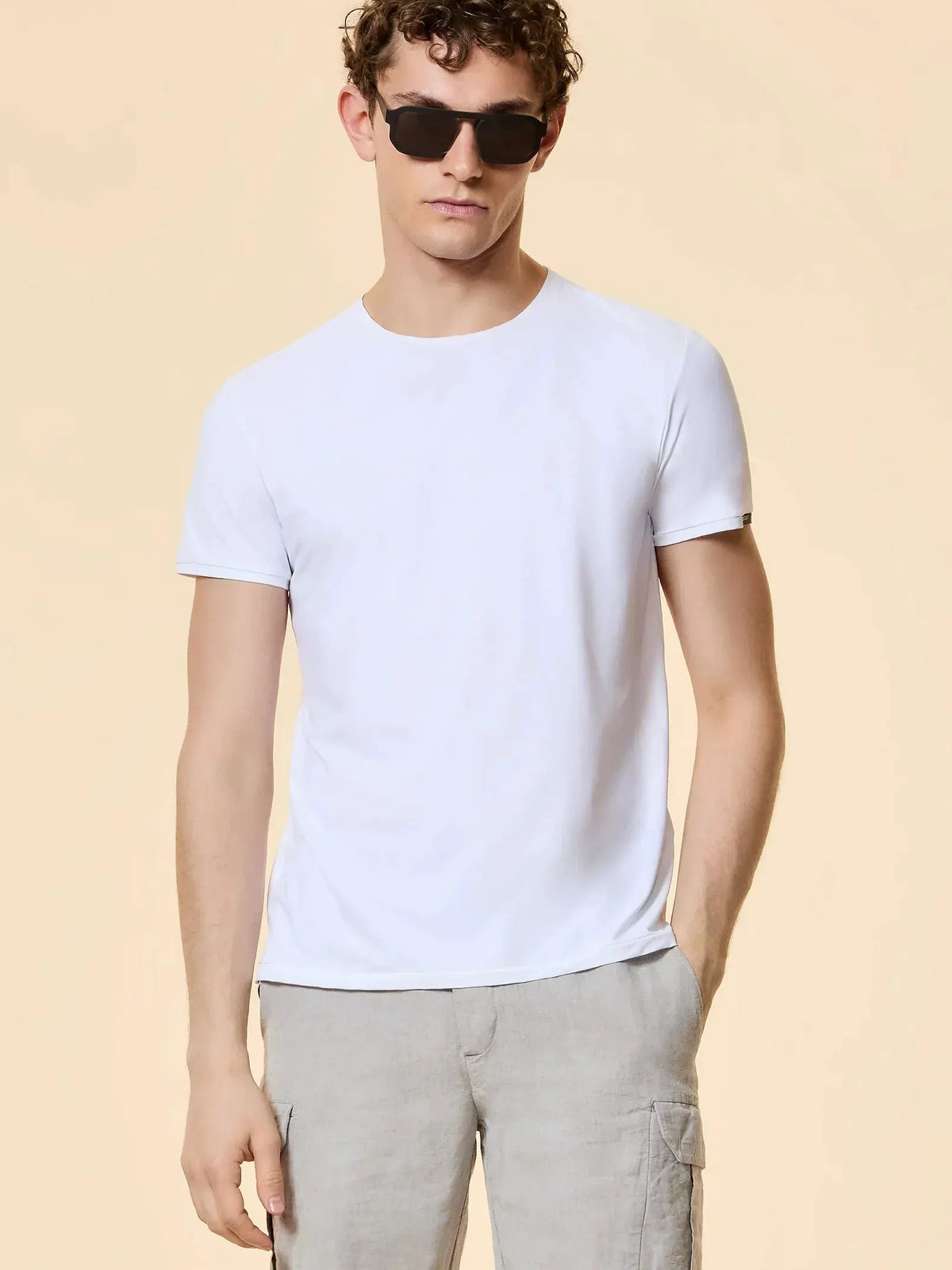 Uomo con maglietta bianca, pantaloni beige in lino e occhiali da sole, sfondo neutro.