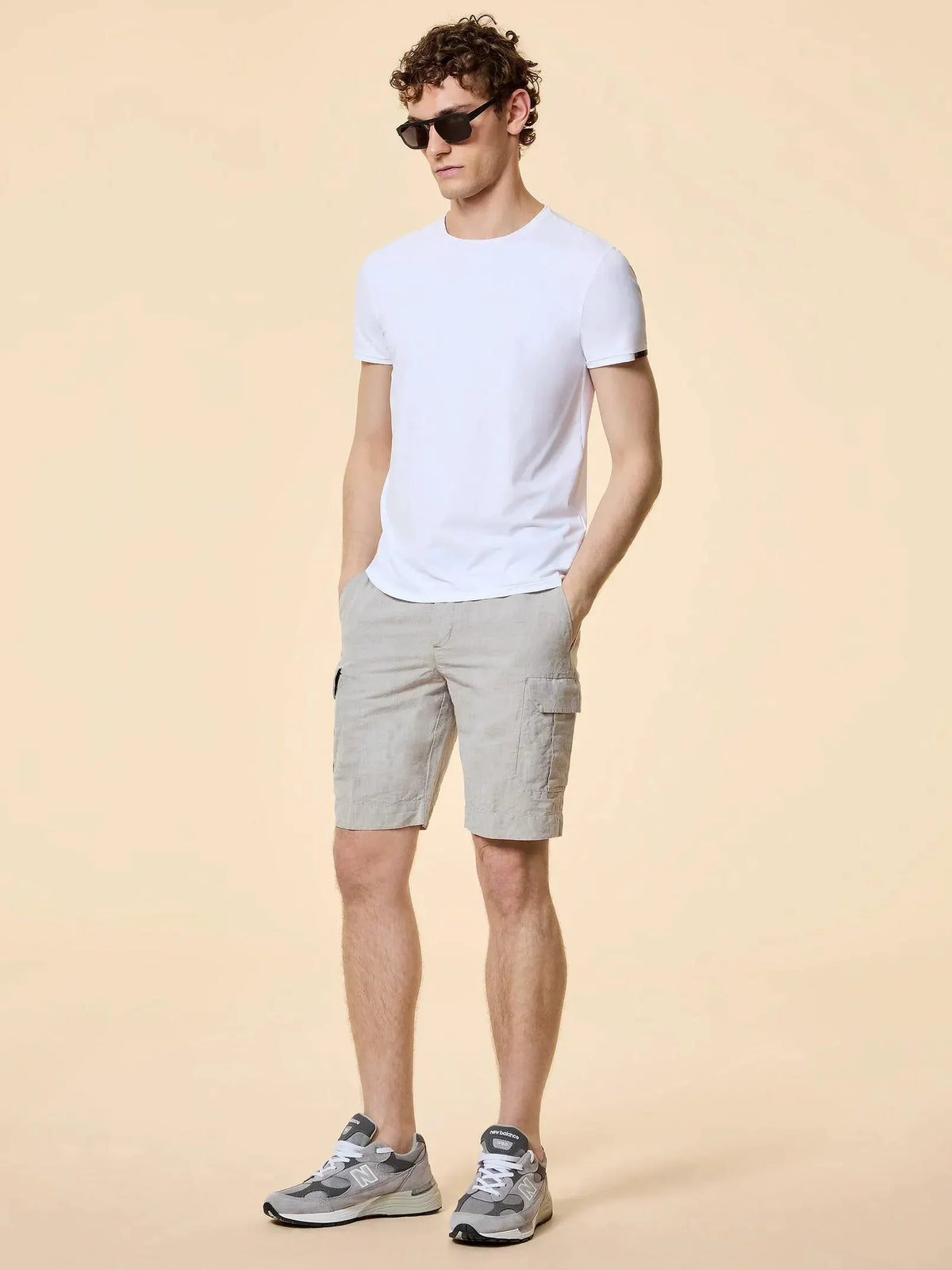 Modello uomo con t-shirt bianca, bermuda beige cargo e sneakers grigie su sfondo neutro.