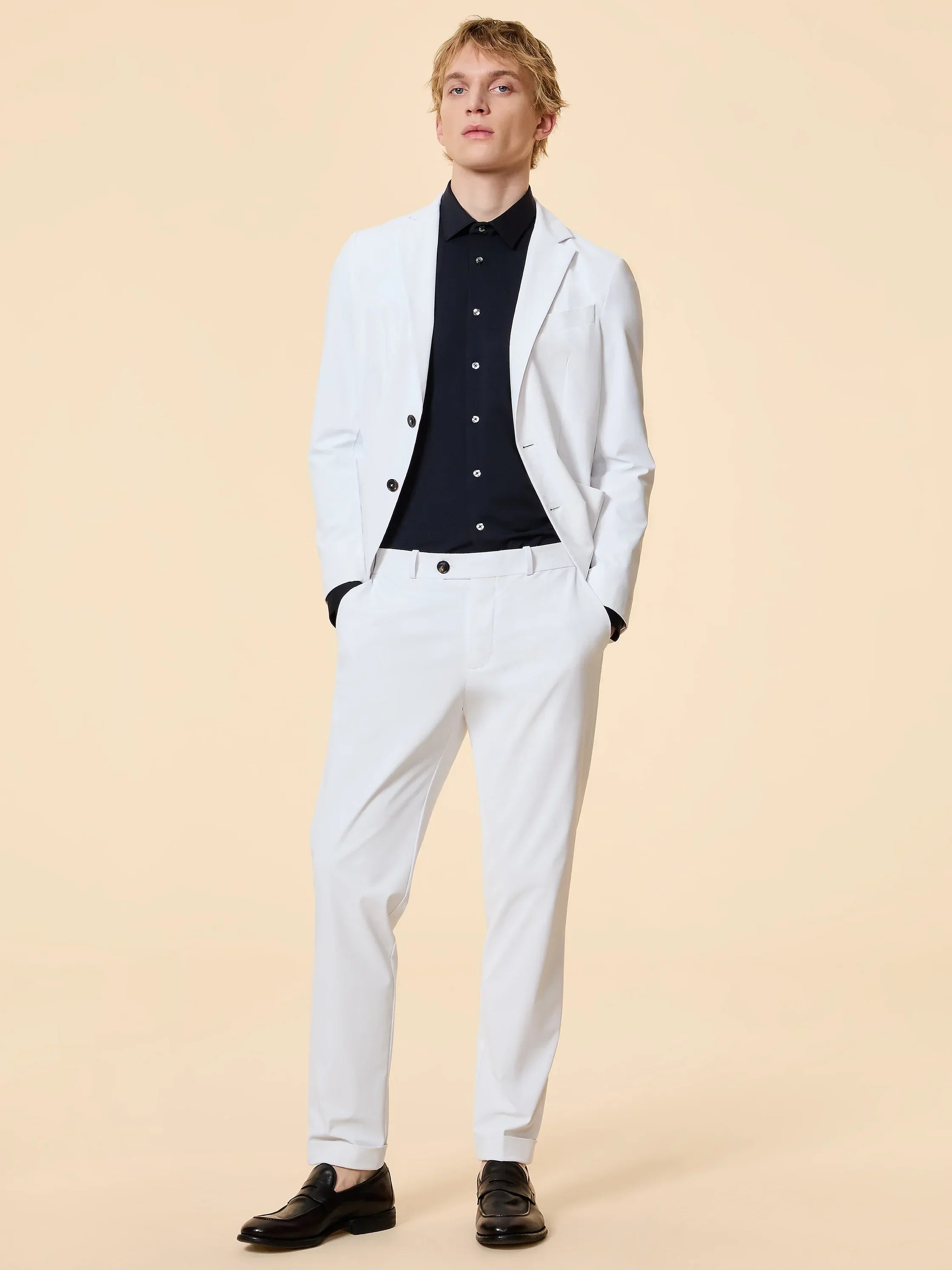 Modello indossa completo uomo elegante bianco con camicia nera e mocassini, su sfondo neutro.