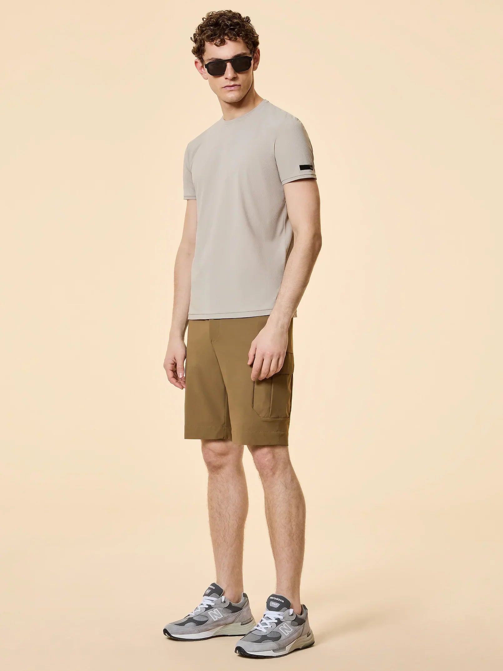 Modello uomo con t-shirt beige, bermuda marrone e sneakers New Balance, stile casual estivo.