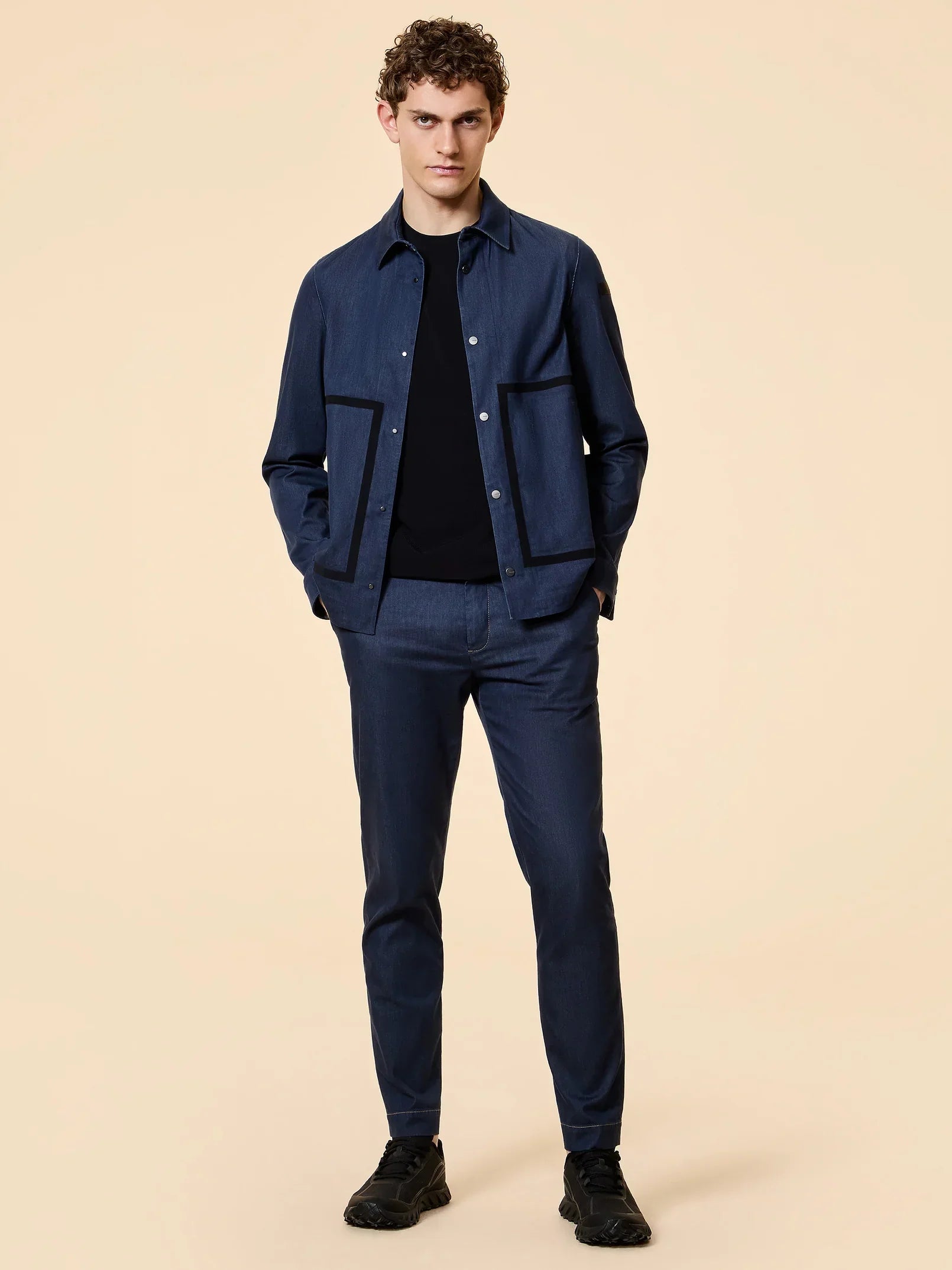 Modello indossa giacca e pantaloni blu stile casual, scarpe nere, sfondo beige.
