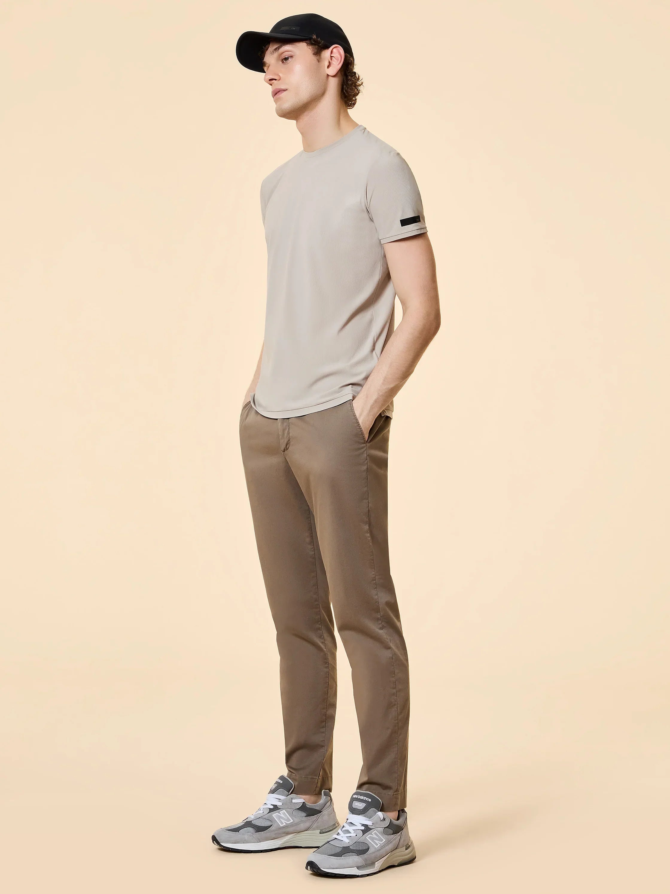 Modello con t-shirt beige, pantaloni slim marroni, sneakers grigie e cappellino nero.