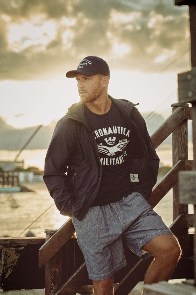 Uomo con giacca blu, t-shirt Aeronautica Militare e cappellino al tramonto su pontile.