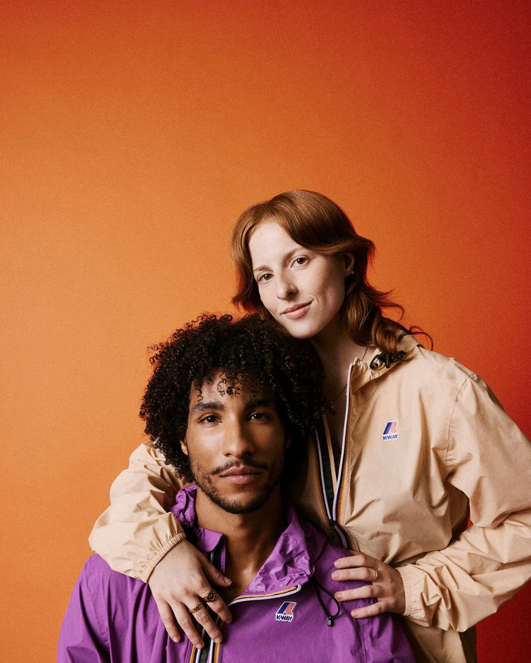 Coppia con giacche K-Way beige e viola su sfondo arancione, moda uomo donna.