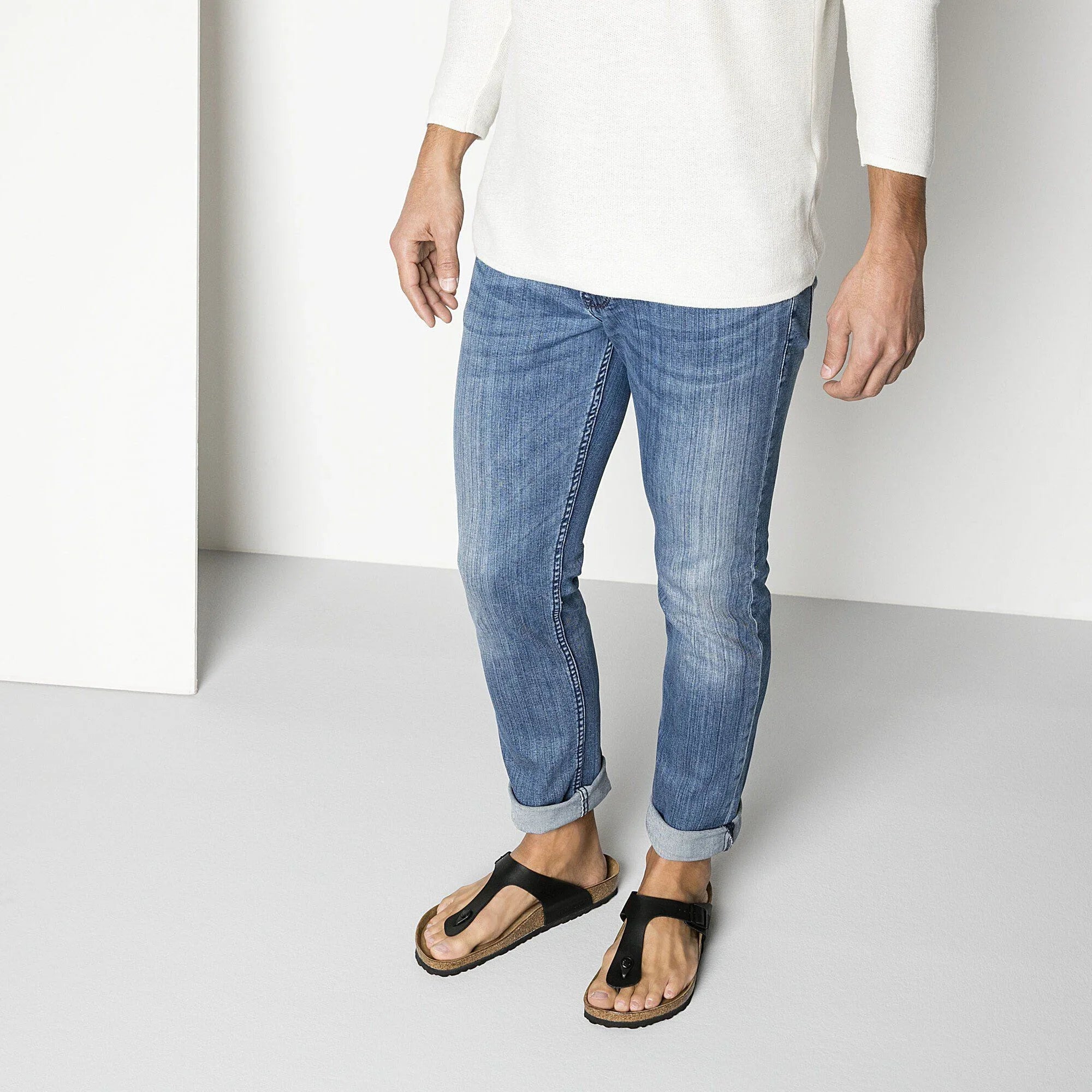 Uomo con jeans slim fit, maglia bianca e sandali infradito neri in stile casual.