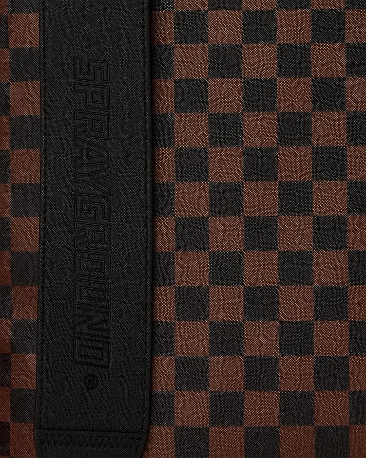 Dettaglio zaino Sprayground con motivo a scacchi marrone e nero e tracolla in pelle nera.