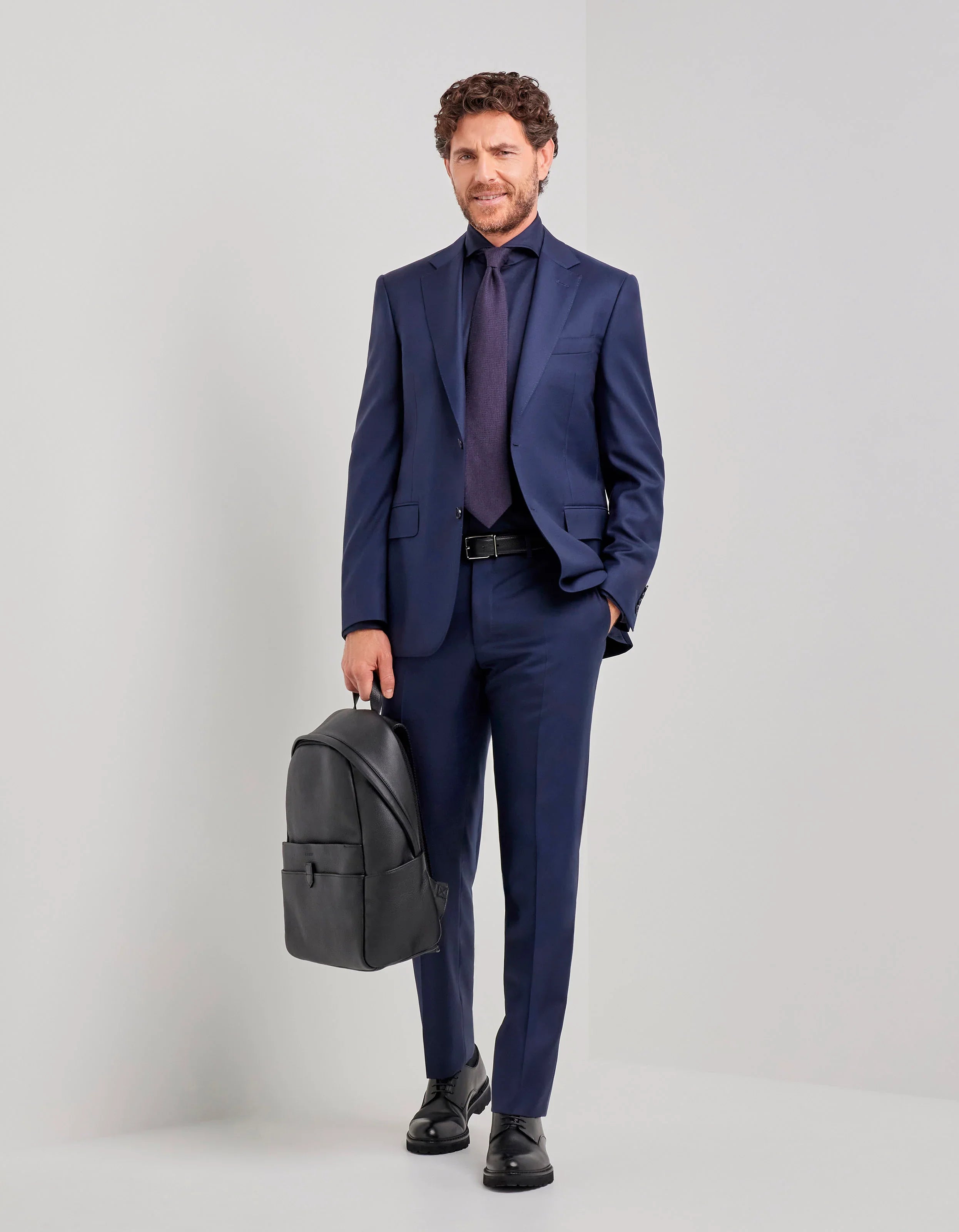 Uomo con completo blu elegante, camicia e cravatta, scarpe nere, zaino nero in mano