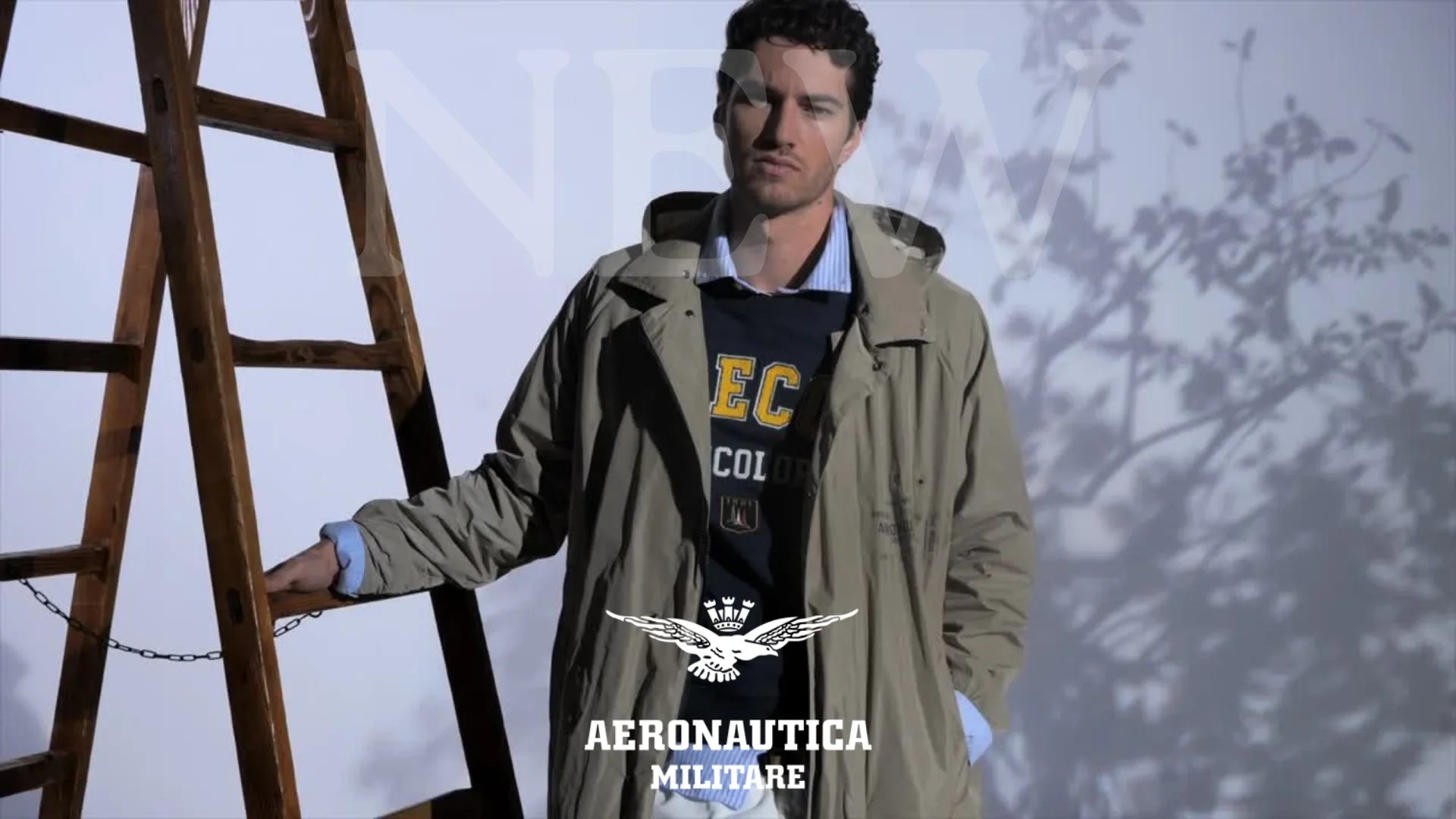 Uomo con trench beige Aeronautica Militare, maglia blu e camicia azzurra, posa vicino a scala in legno.