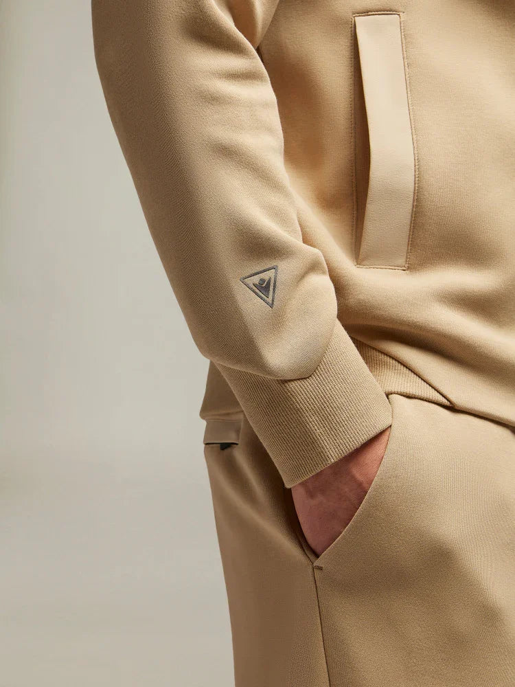 Completo tuta beige uomo con dettaglio logo sulla manica e mano in tasca, stile casual.