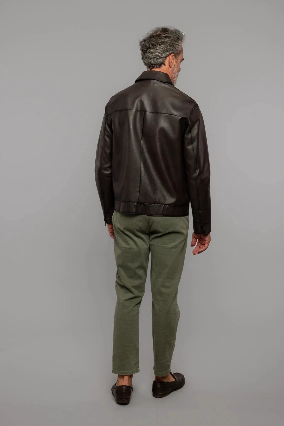 Uomo di spalle con giacca in pelle marrone, pantaloni verdi e mocassini, sfondo neutro.