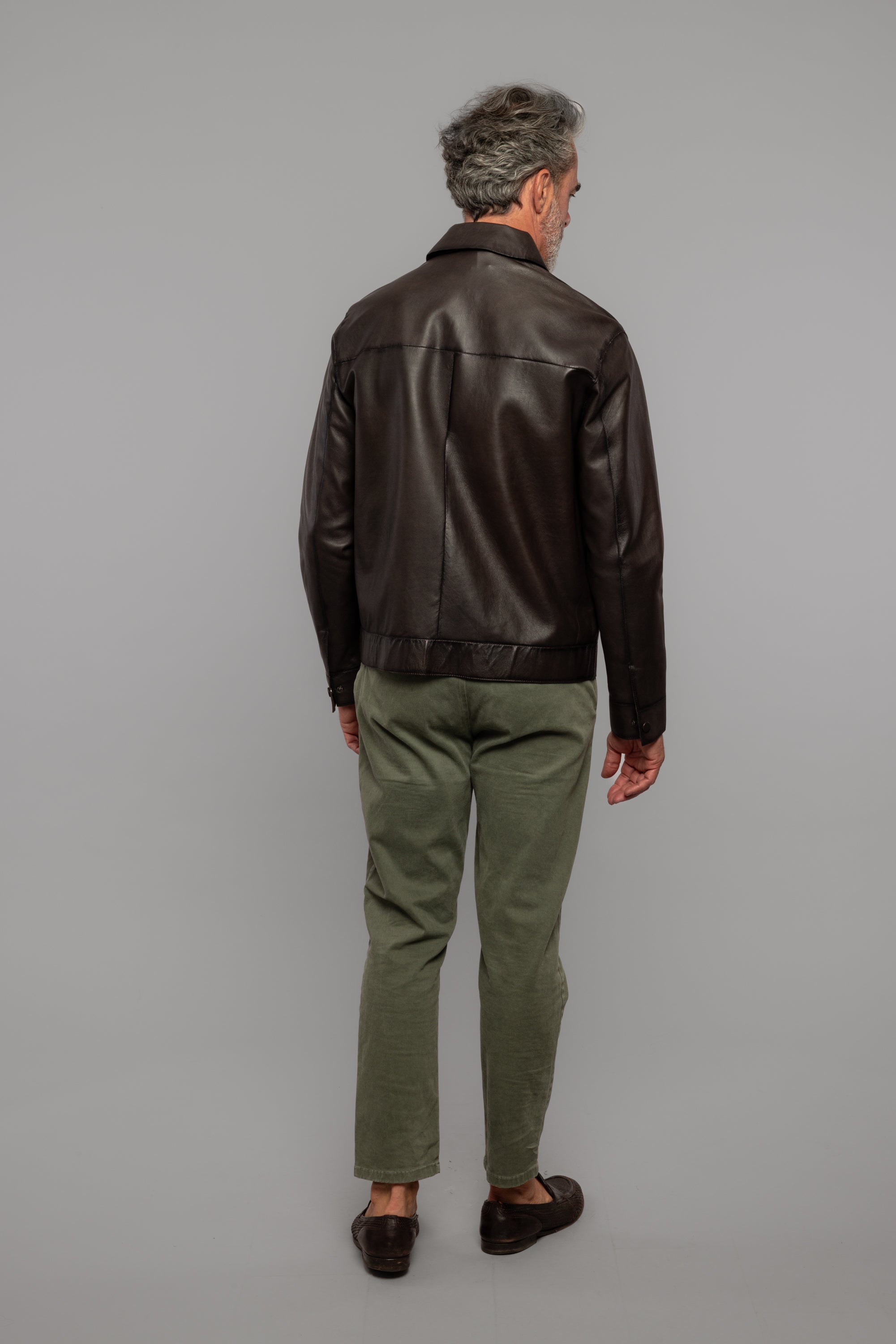 Uomo di spalle con giacca in pelle marrone, pantaloni verdi e mocassini, sfondo neutro.