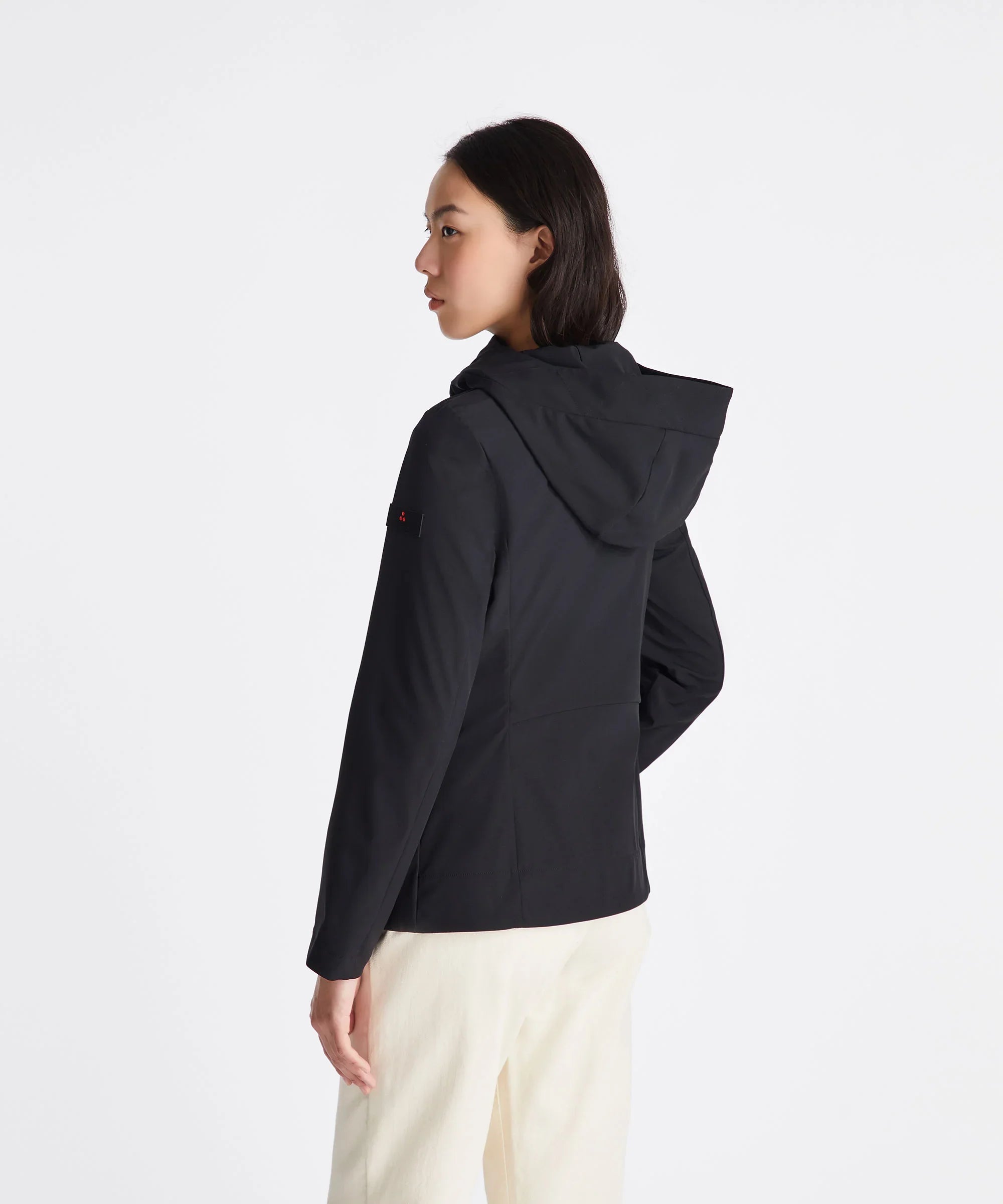 Donna di spalle con giacca nera con cappuccio e pantaloni chiari su sfondo bianco.