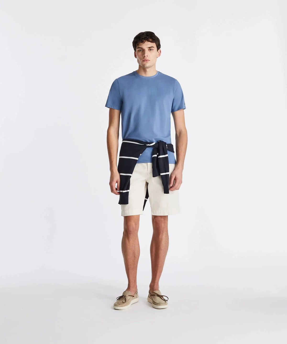Uomo con t-shirt azzurra, bermuda bianchi, maglione a righe in vita e mocassini beige