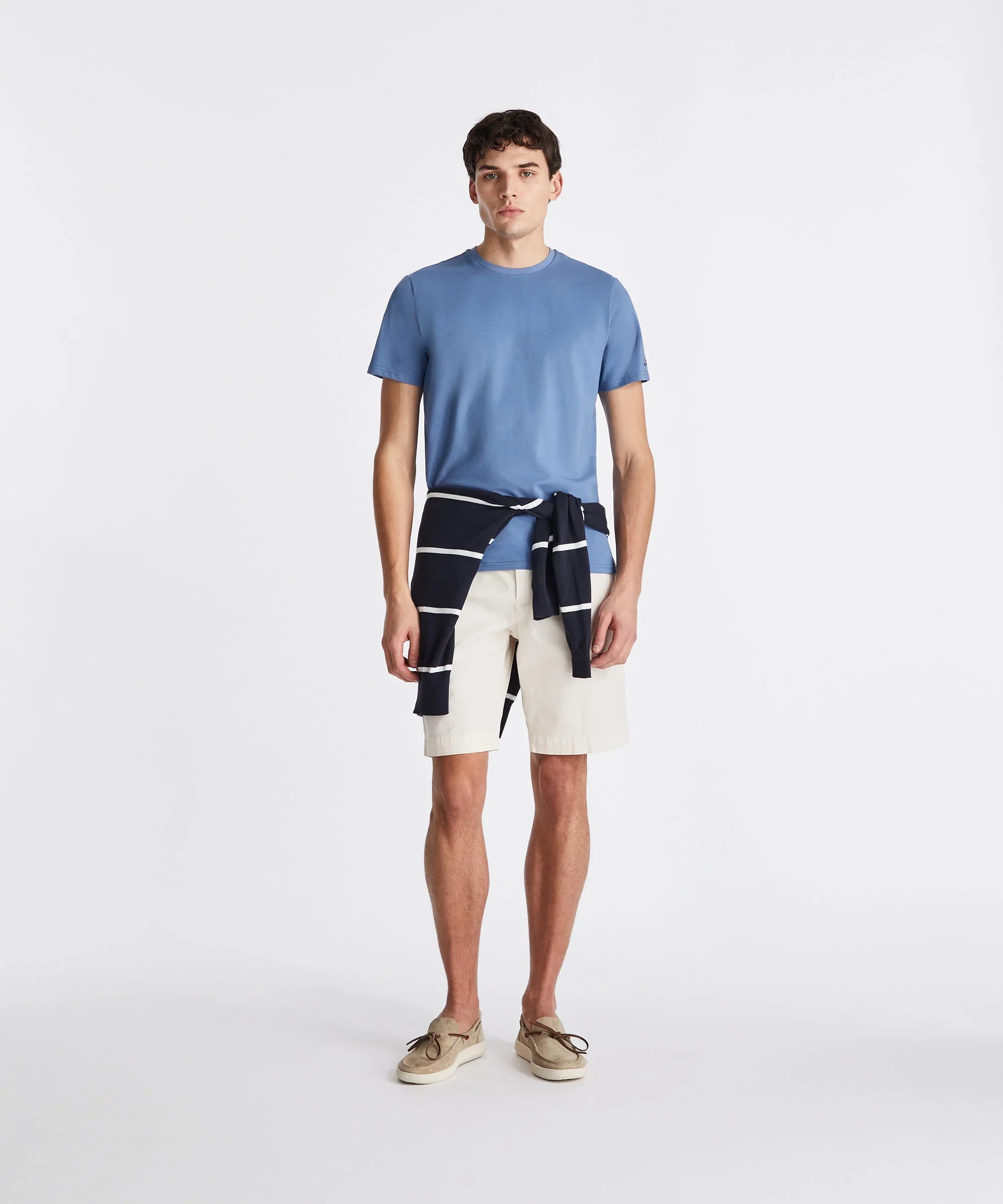 Uomo con t-shirt azzurra, bermuda bianchi, maglione a righe in vita e mocassini beige