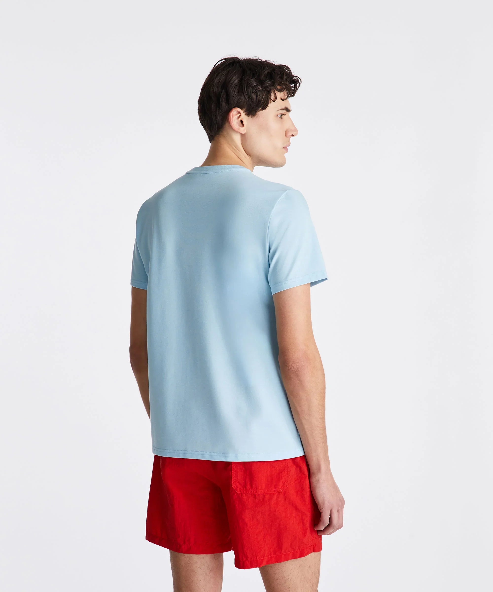 Uomo di profilo con t-shirt celeste e shorts rossi su sfondo bianco, moda estiva casual.