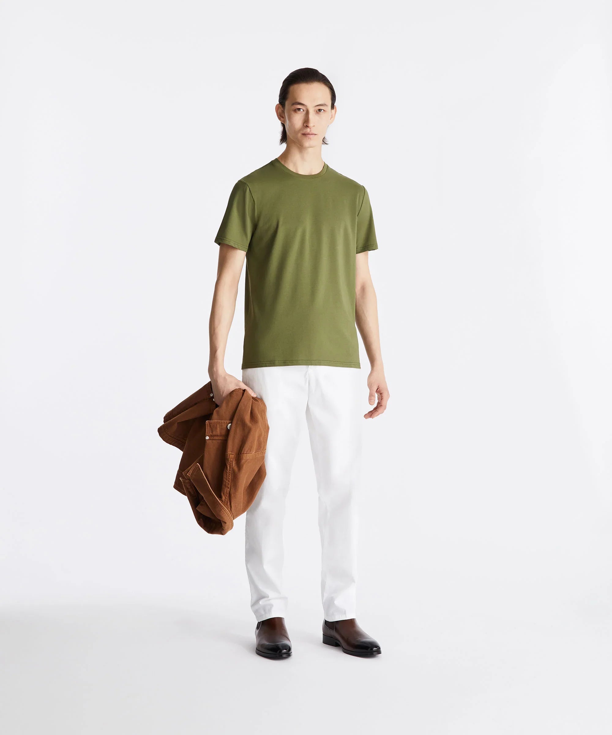 Uomo con t-shirt verde, pantaloni bianchi, giacca marrone e stivaletti scuri su sfondo bianco