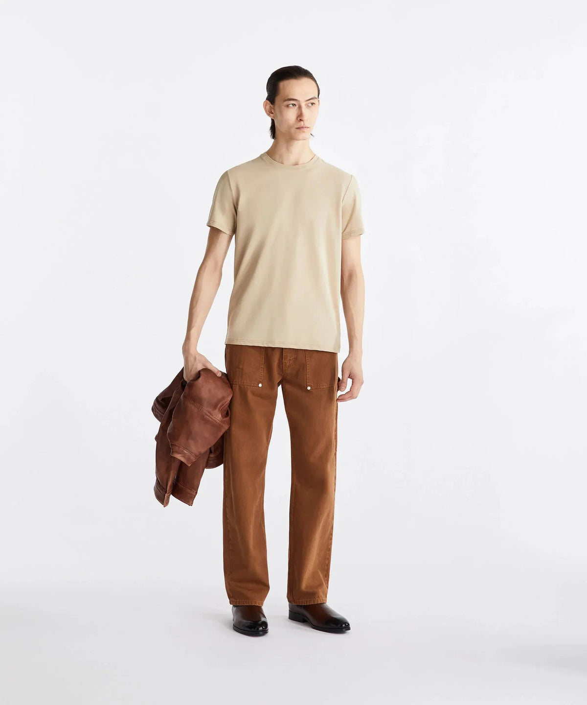 Modello indossa t-shirt beige e pantaloni marroni, con giacca marrone in mano, su sfondo bianco.