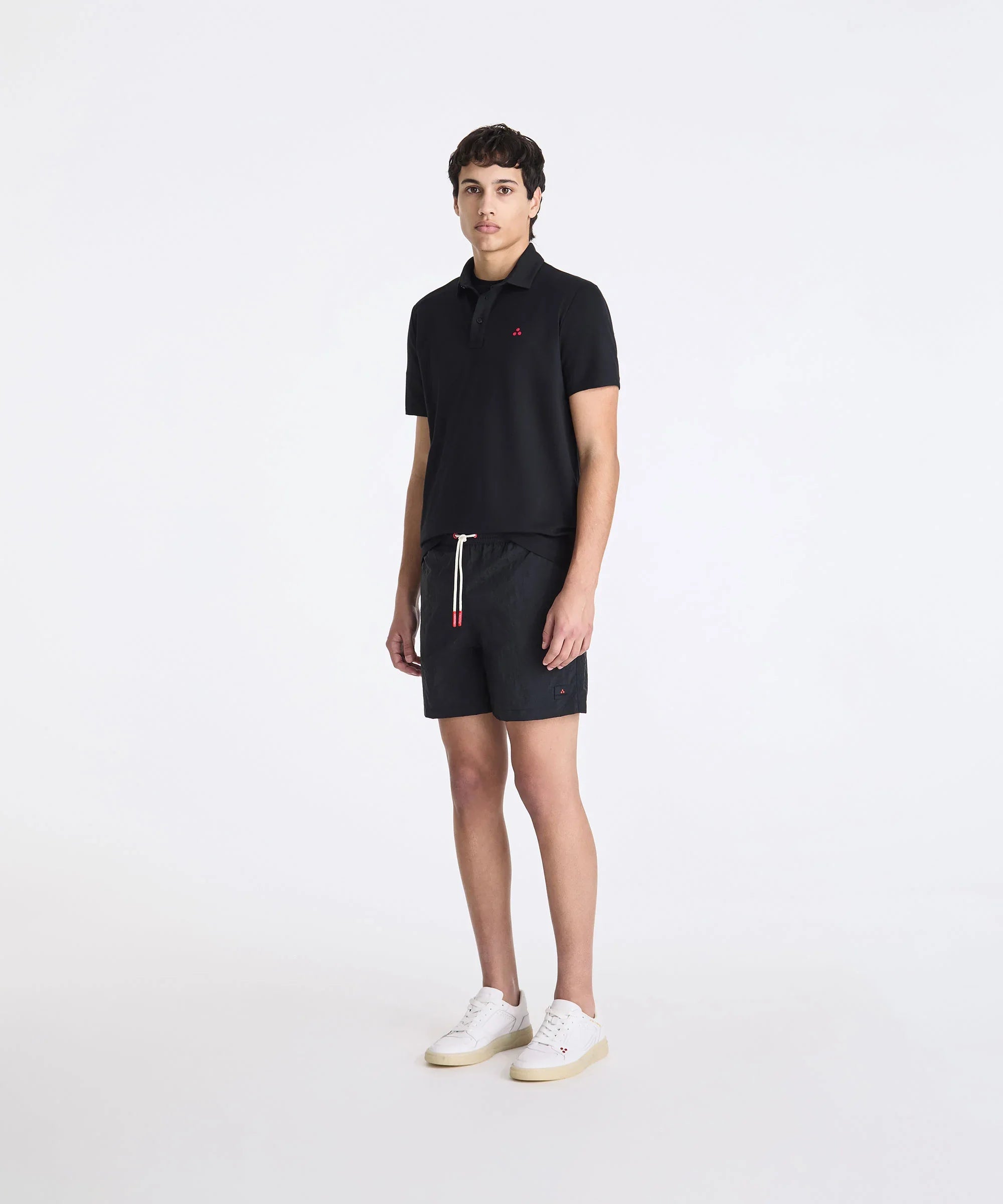 Modello indossa polo nera uomo, bermuda neri e sneakers bianche su sfondo chiaro