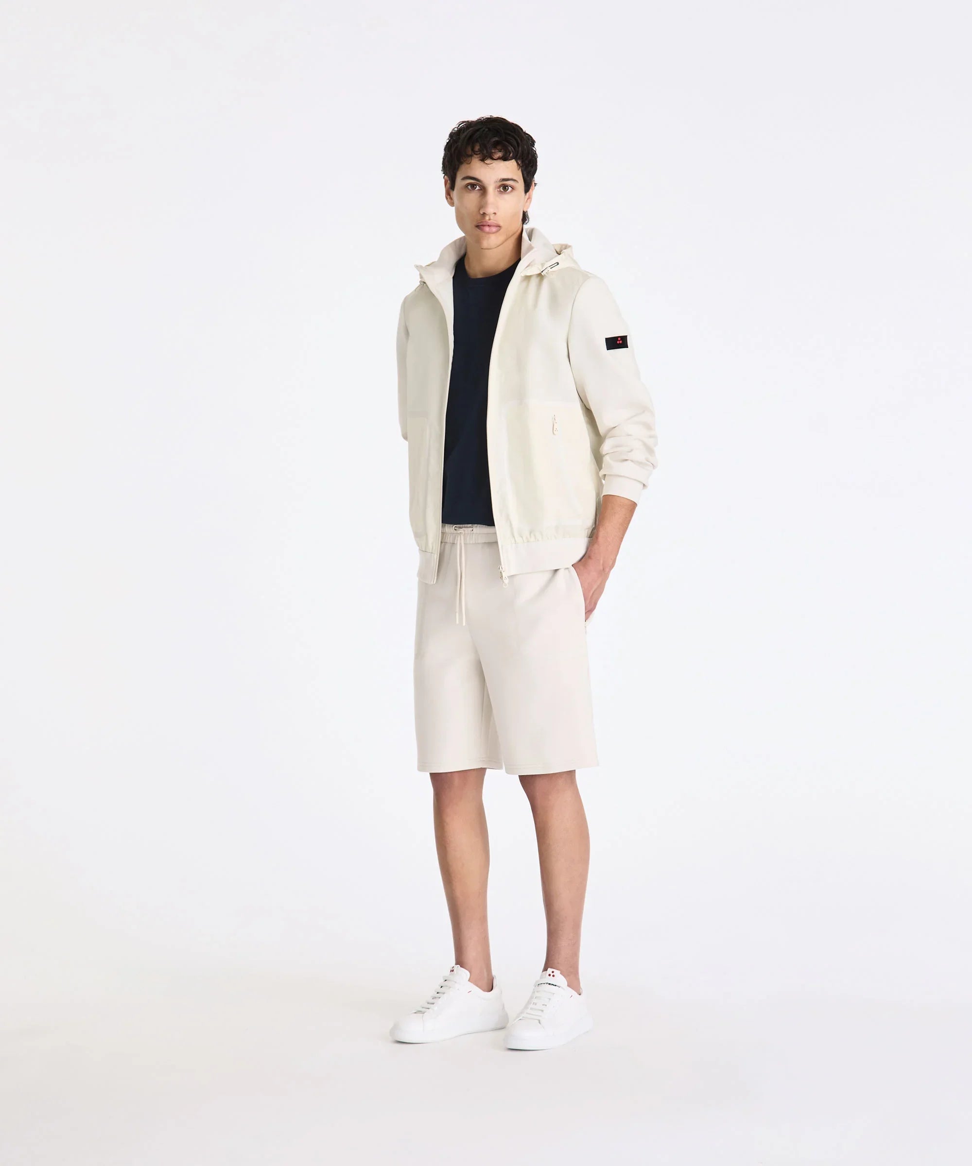 Modello indossa giacca con cappuccio beige, t-shirt nera, bermuda beige e sneakers bianche uomo.
