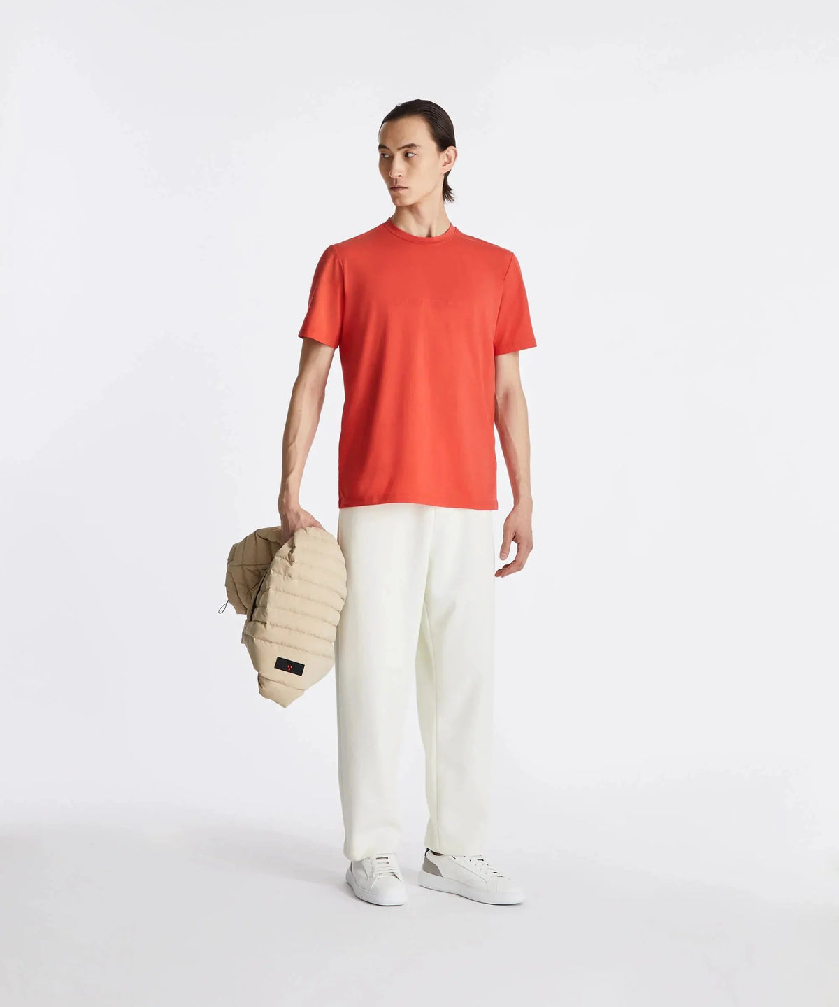 Uomo con t-shirt arancione, pantaloni bianchi, scarpe bianche e piumino beige in mano