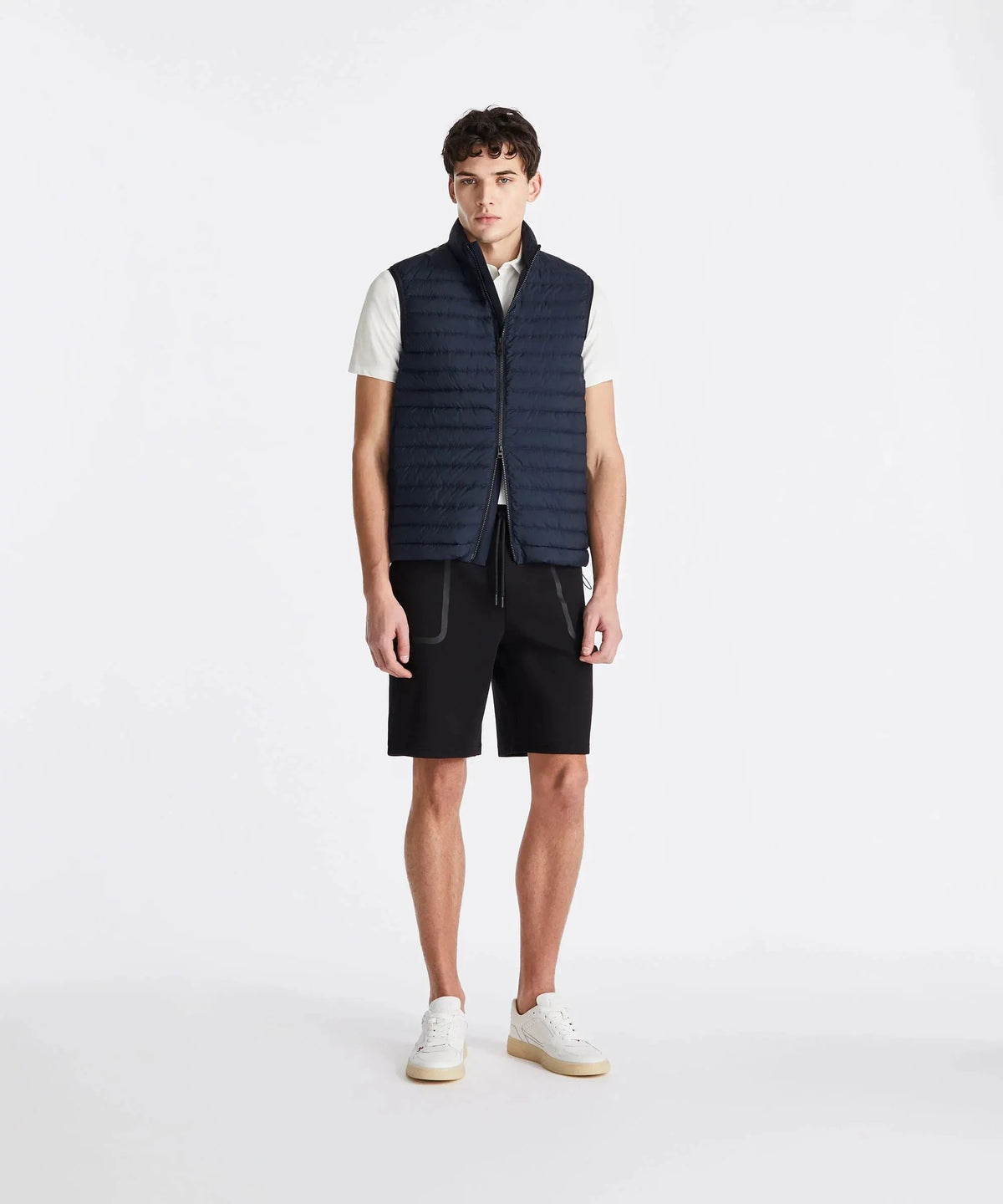 Modello uomo con gilet trapuntato blu, polo bianca, bermuda neri e sneakers bianche
