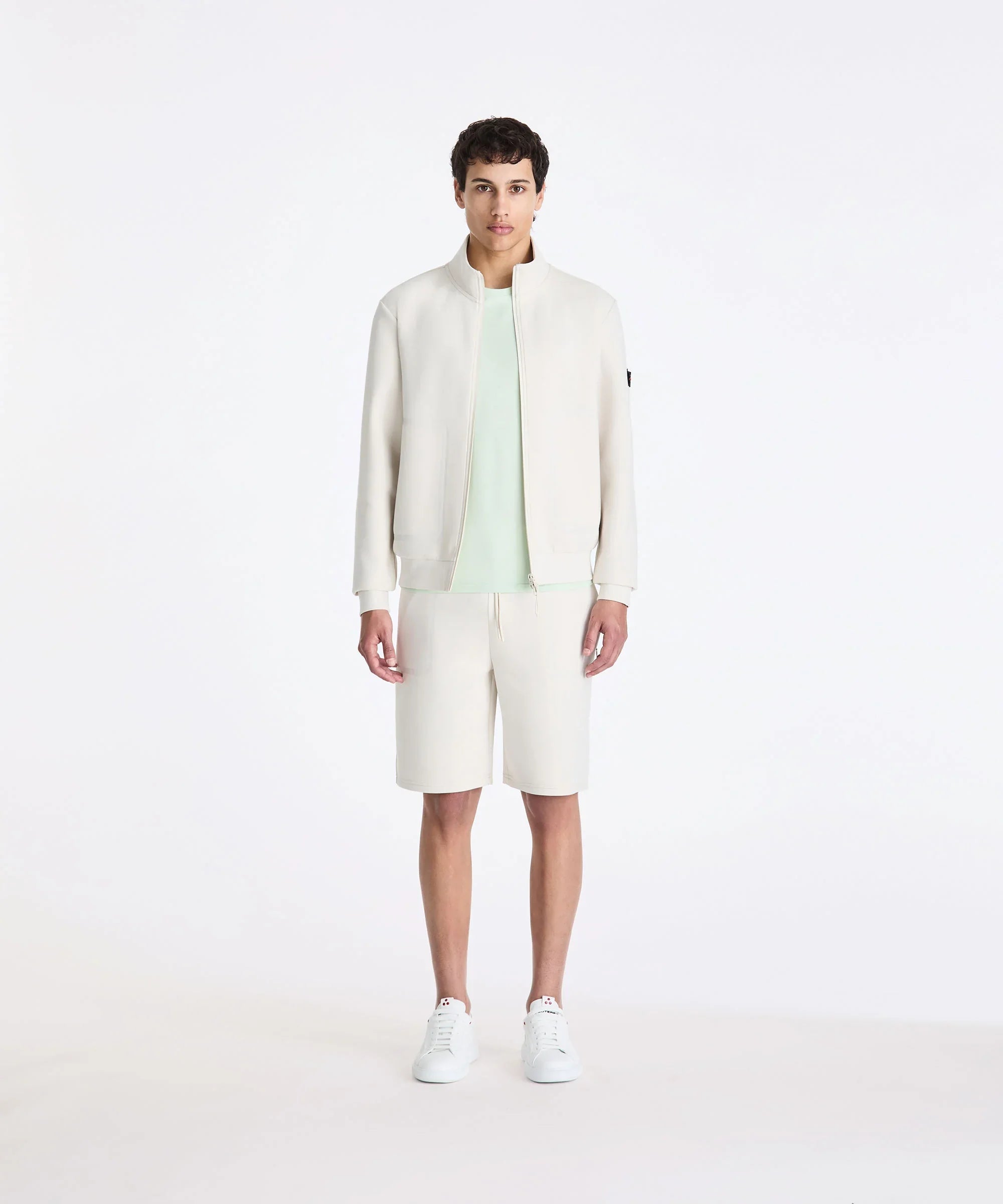 Modello uomo con giacca e bermuda color panna, t-shirt verde chiaro e sneakers bianche