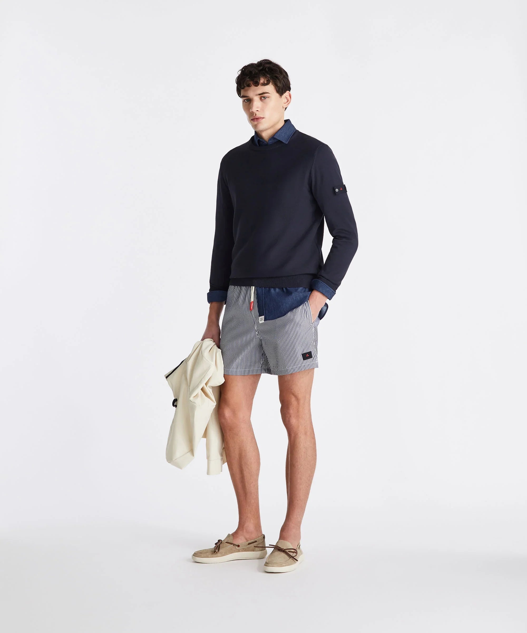 Modello uomo con pullover blu, camicia jeans, bermuda rigati e mocassini beige, stile casual.