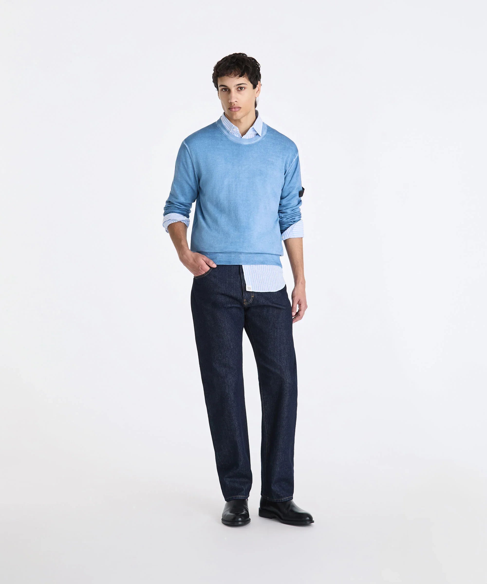 Modello uomo con maglione azzurro, camicia a righe e jeans scuri su sfondo bianco.