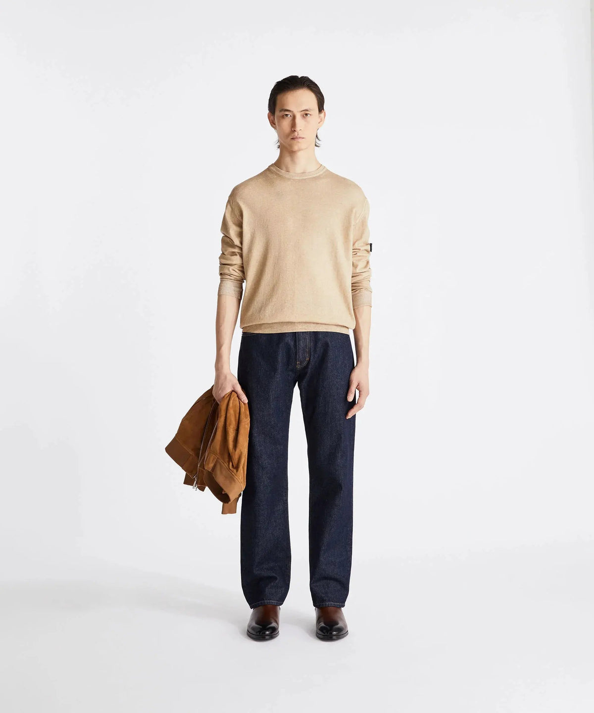 Modello uomo con maglione beige, jeans blu, scarpe marroni, giacca in suede in mano, sfondo bianco.