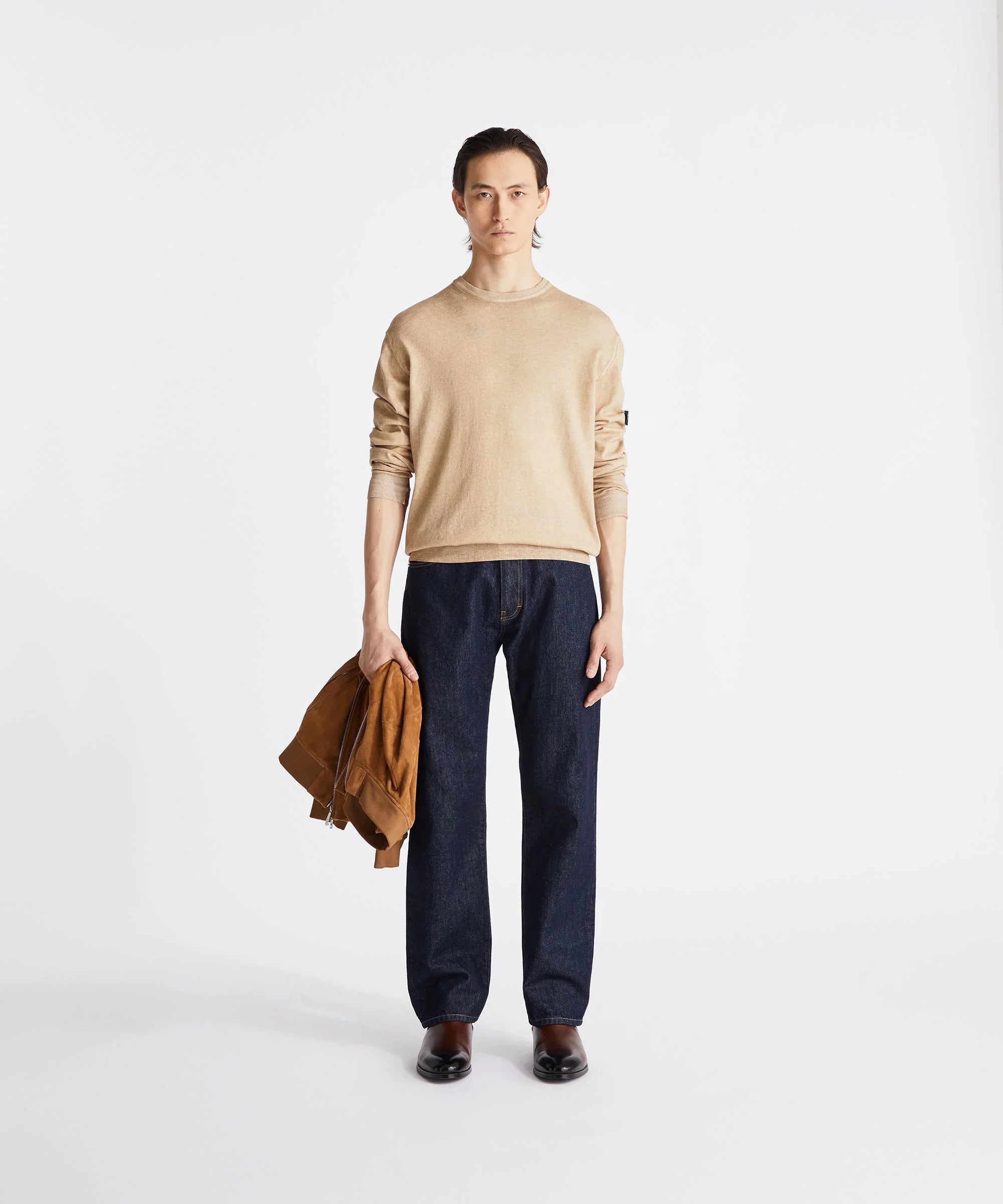 Modello uomo con maglione beige, jeans blu, scarpe marroni, giacca in suede in mano, sfondo bianco.