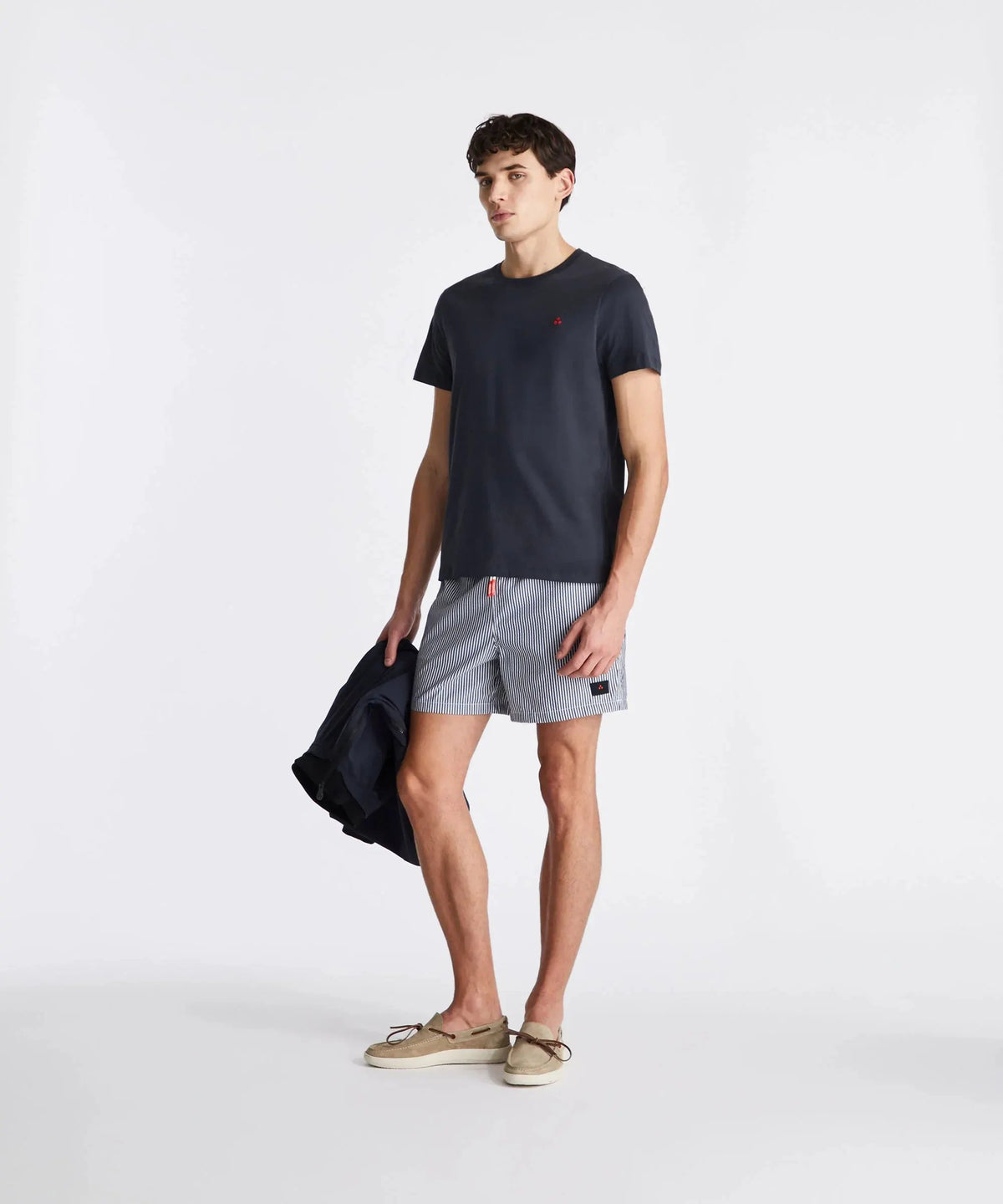 Modello uomo con t-shirt blu, bermuda rigati, mocassini beige e giacca in mano.