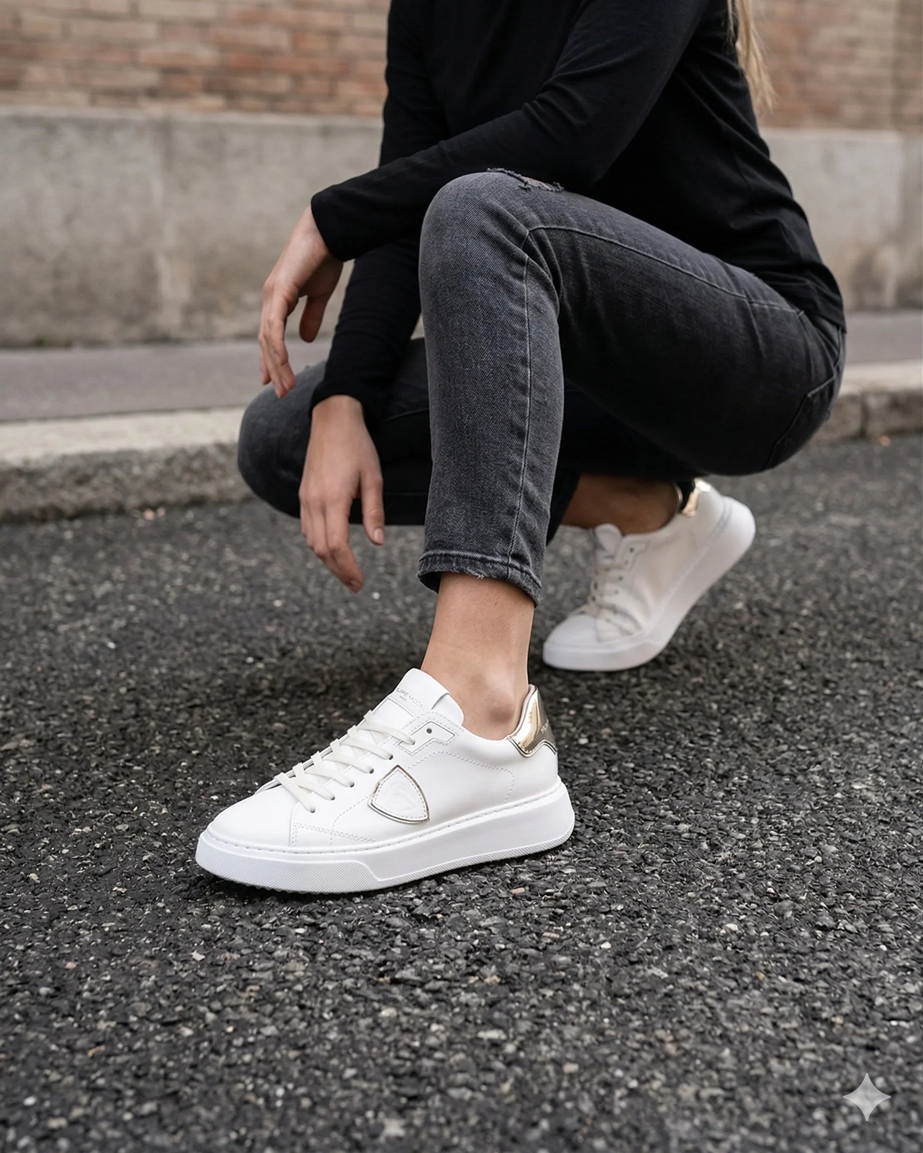 Sneakers bianche da donna con dettagli dorati, abbinate a jeans neri e maglia nera, indossate all'aperto.
