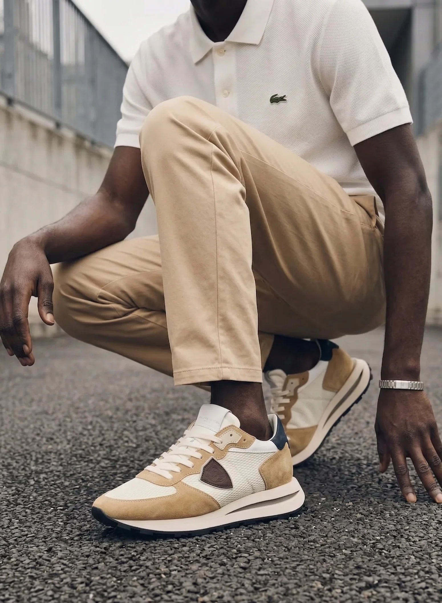 Sneakers beige e bianche da uomo, pantaloni chino beige e polo bianca su sfondo urbano.
