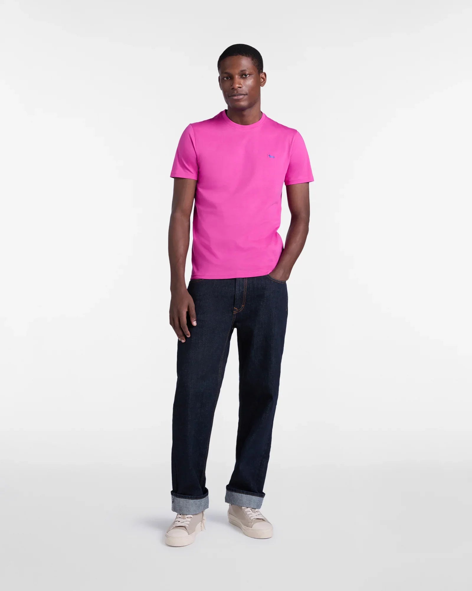 Modello uomo indossa t-shirt fucsia a maniche corte, jeans blu e sneakers beige su sfondo bianco.