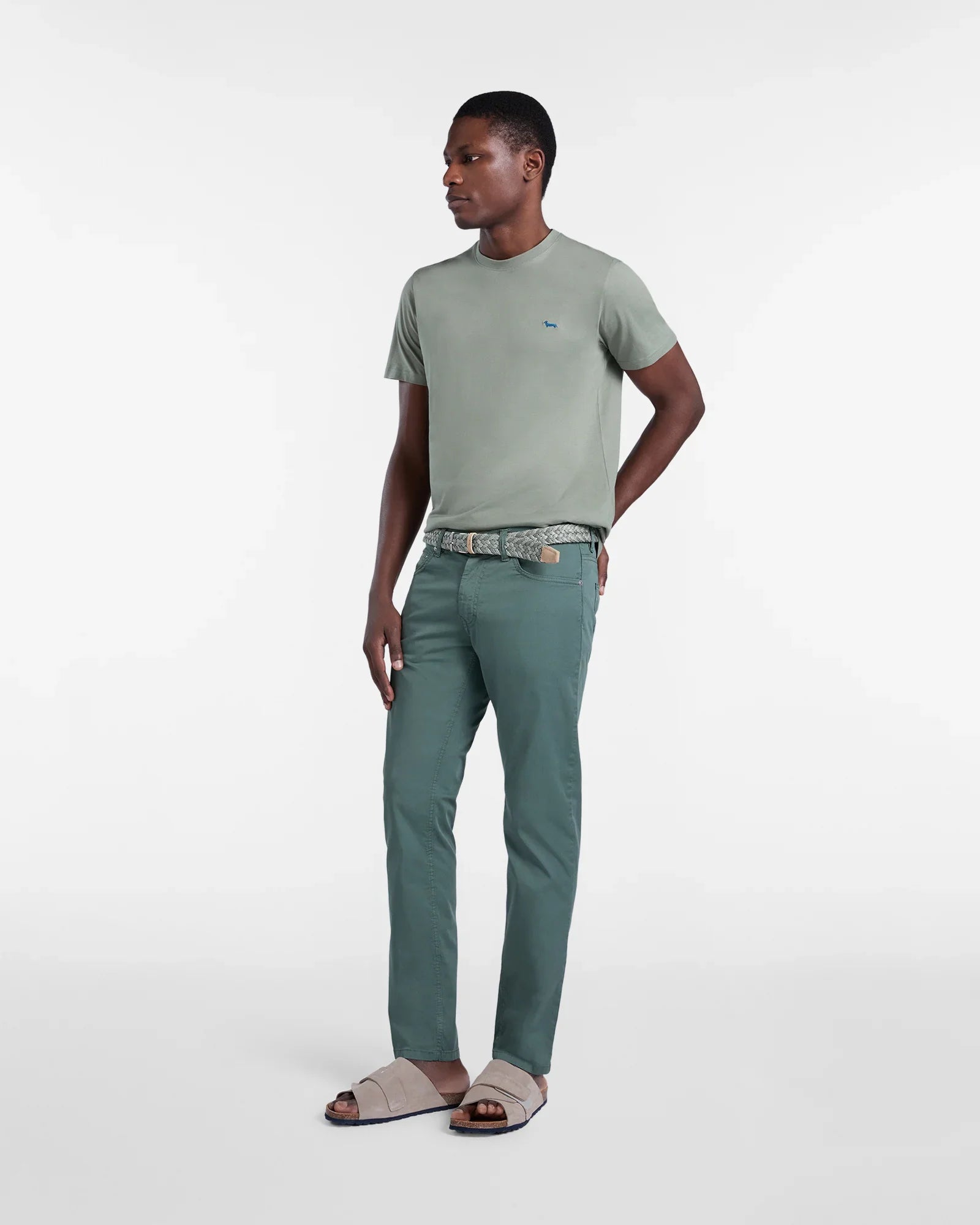 Modello uomo con t-shirt verde salvia, pantaloni verde, cintura intrecciata e sandali beige.
