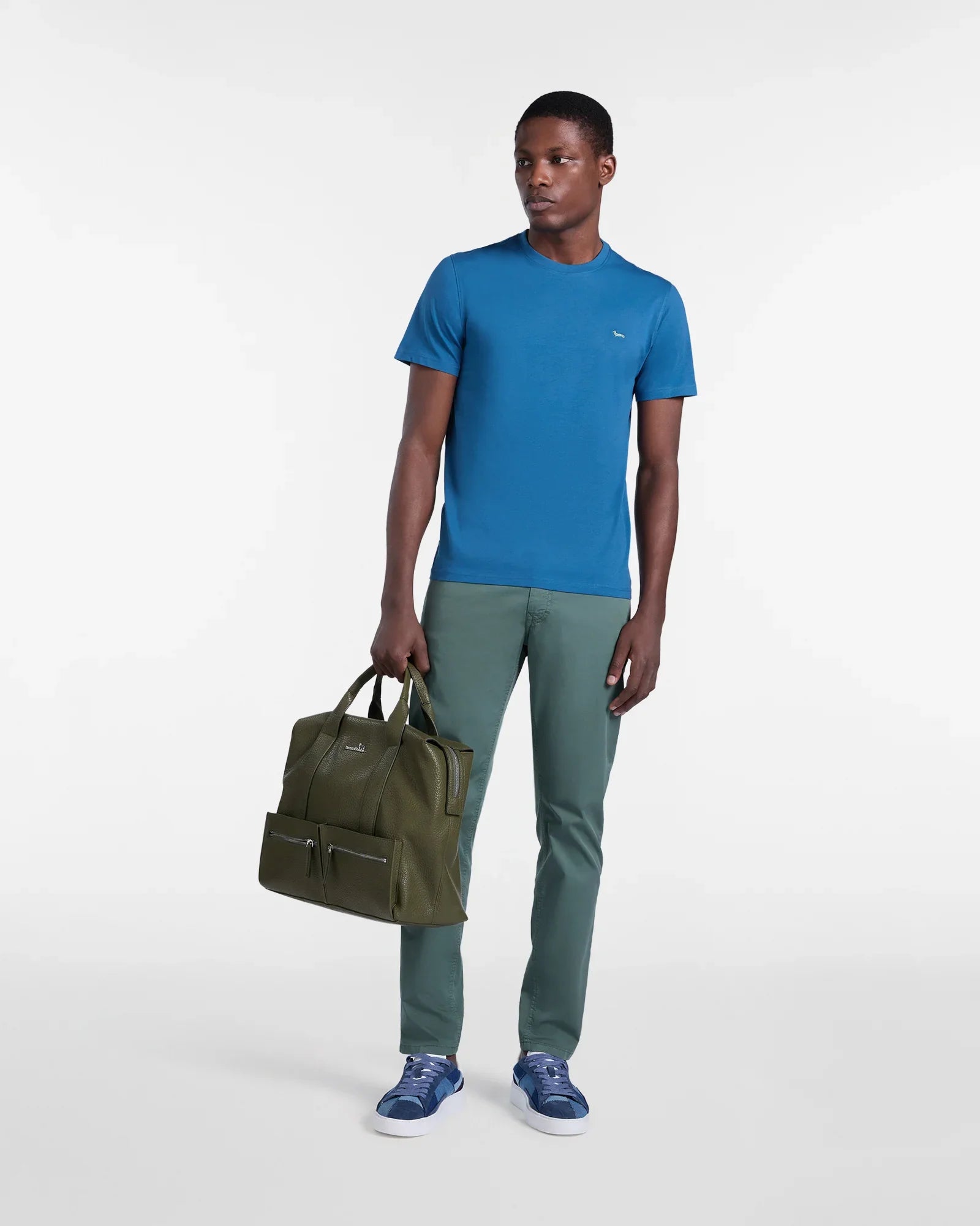 Modello uomo con t-shirt blu, pantaloni verdi, sneakers blu e borsa verde elegante.