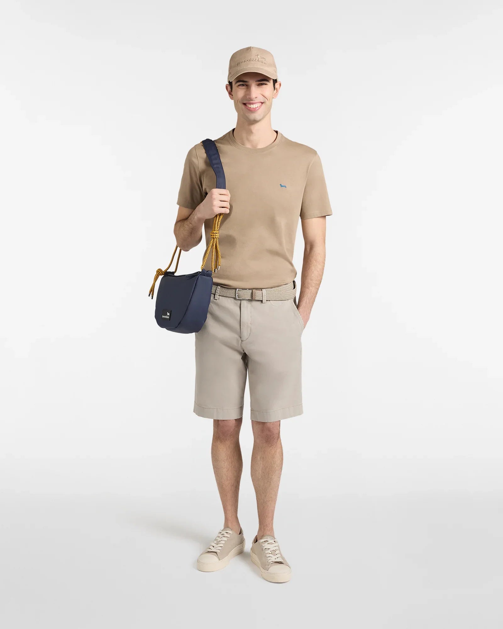 Uomo con t-shirt beige, bermuda chiari, sneakers e borsa blu a tracolla, look casual estivo.
