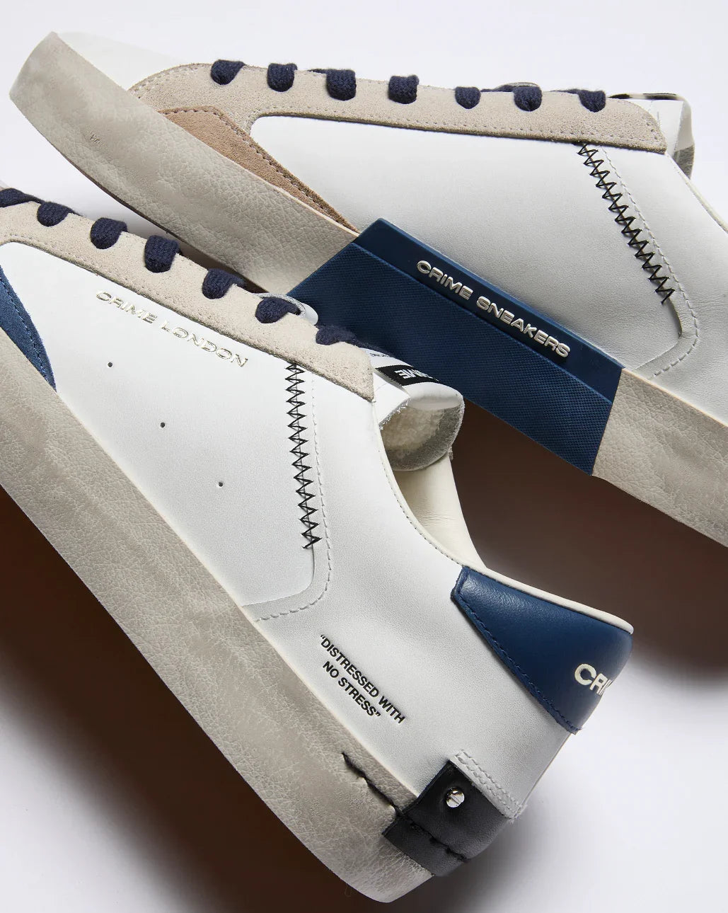Sneakers bianche da uomo con dettagli blu, beige e neri, suola effetto used, marchio Crime London visibile.
