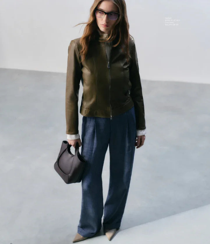 Donna con giacca di pelle verde, pantaloni eleganti blu, borsa scura e occhiali da vista
