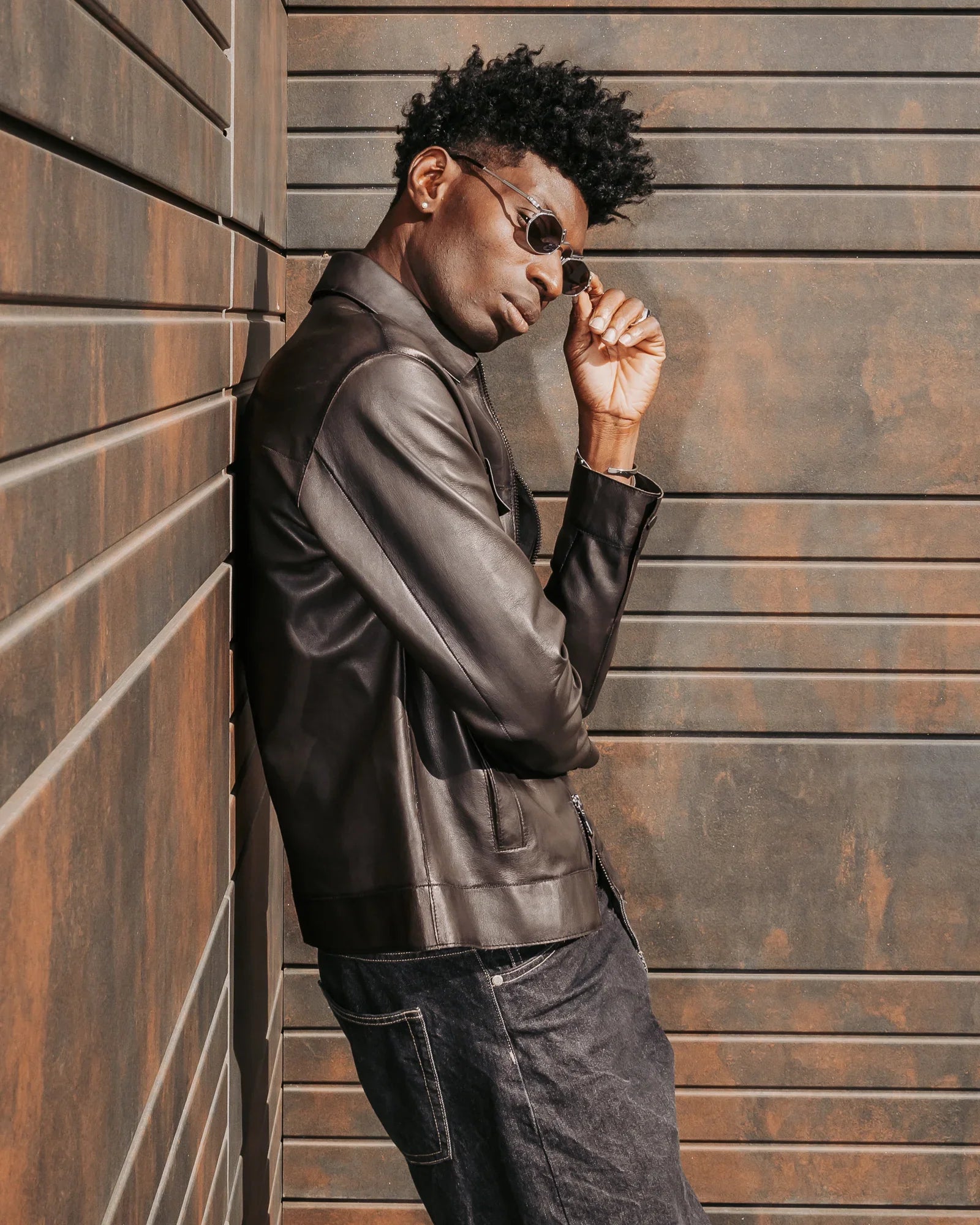 Uomo con giacca di pelle marrone e jeans, occhiali da sole, posa su sfondo industriale