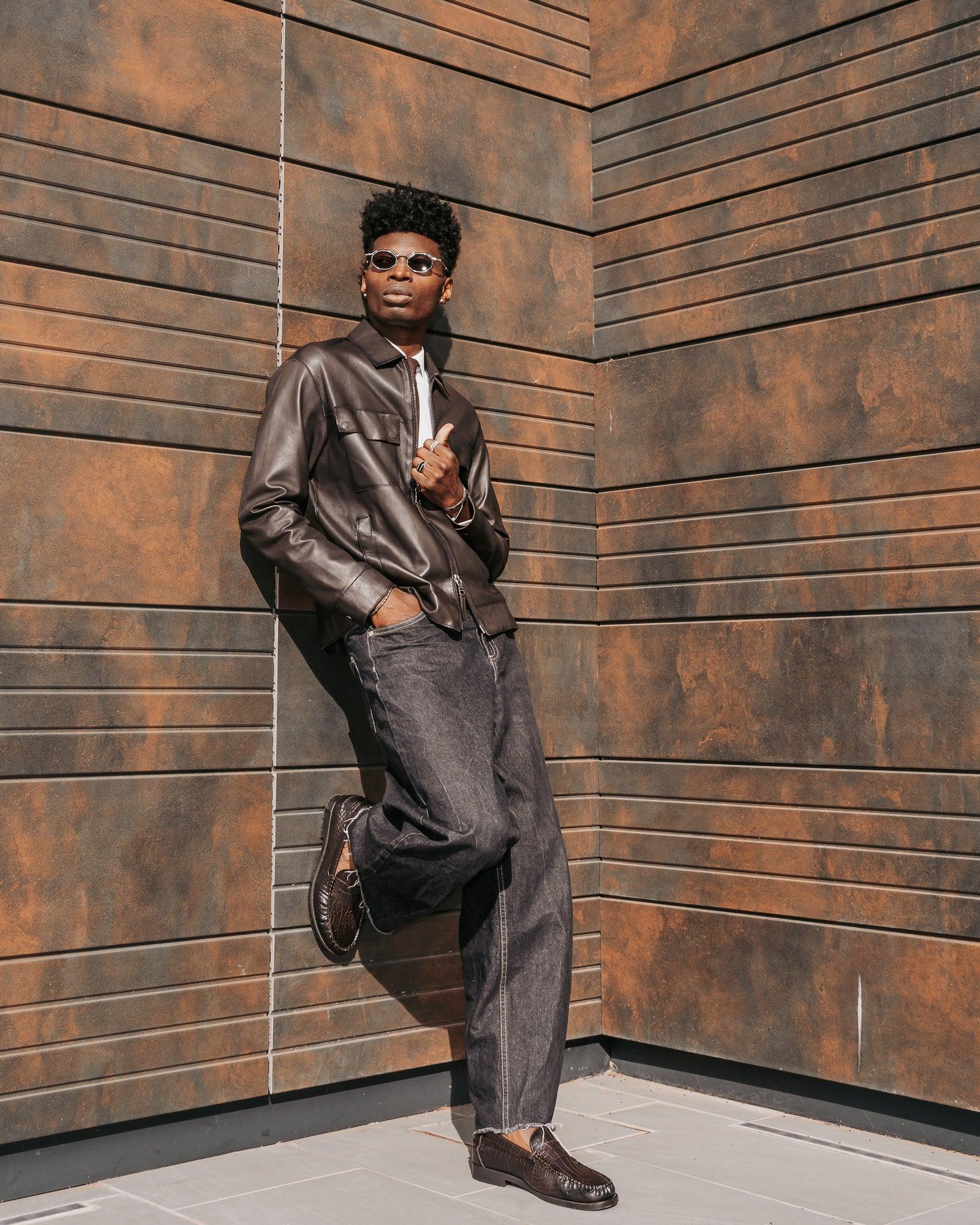Uomo con giacca in pelle marrone, jeans scuri e mocassini, posa outdoor moderna, moda uomo elegante.