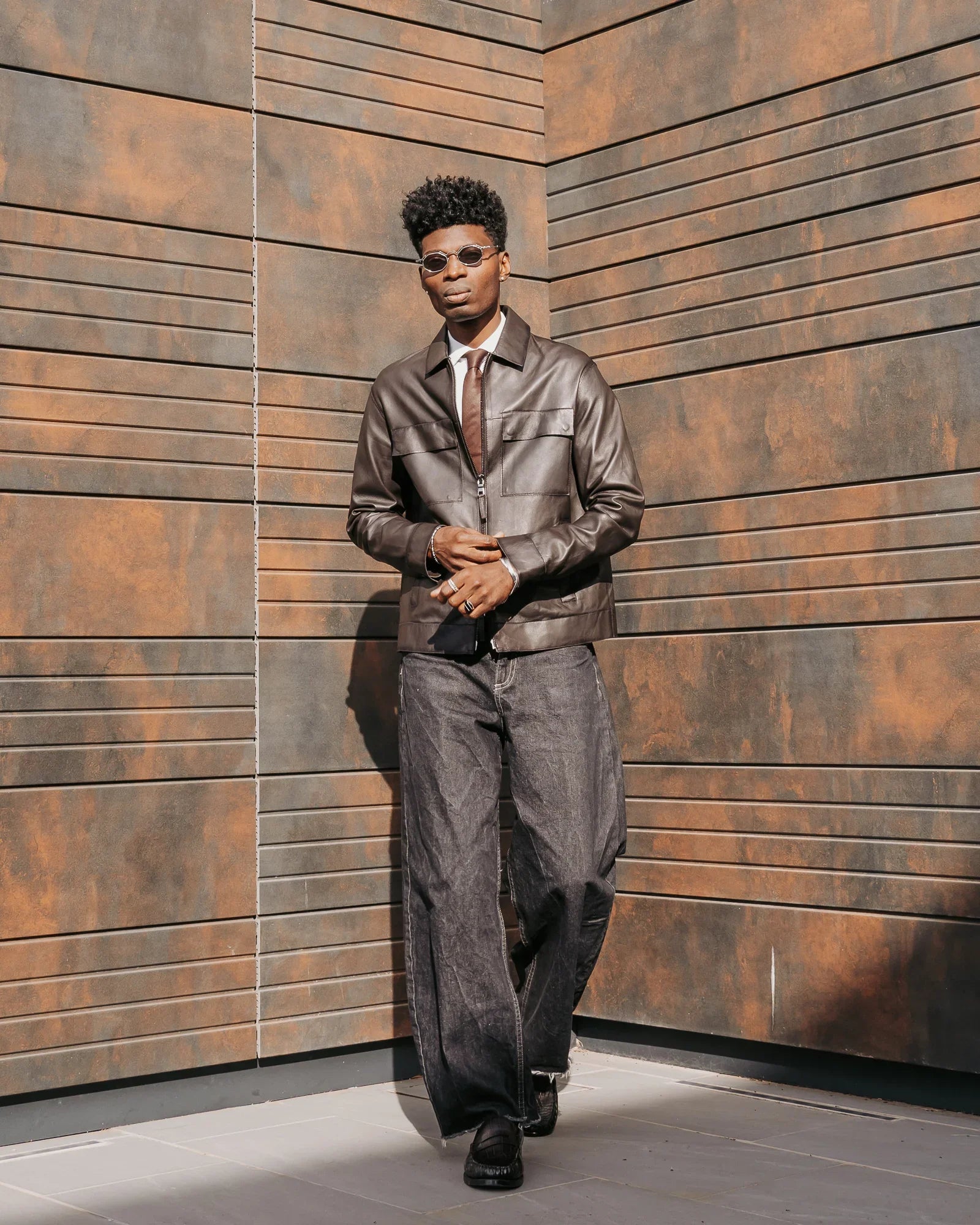 Uomo con giacca in pelle marrone, pantaloni larghi grigi e occhiali da sole, moda uomo elegante.