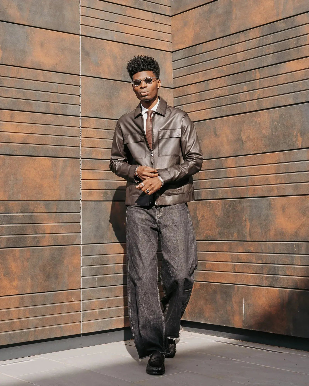 Uomo con giacca in pelle marrone, pantaloni larghi grigi e occhiali da sole, moda uomo elegante.